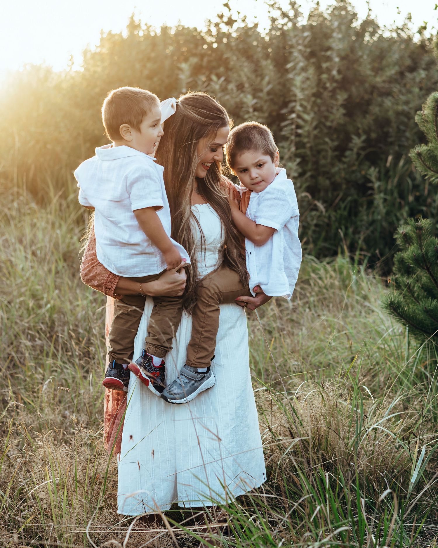 Three people embrace tenderly in a sunlit meadow with tall grass and trees at golden hour.