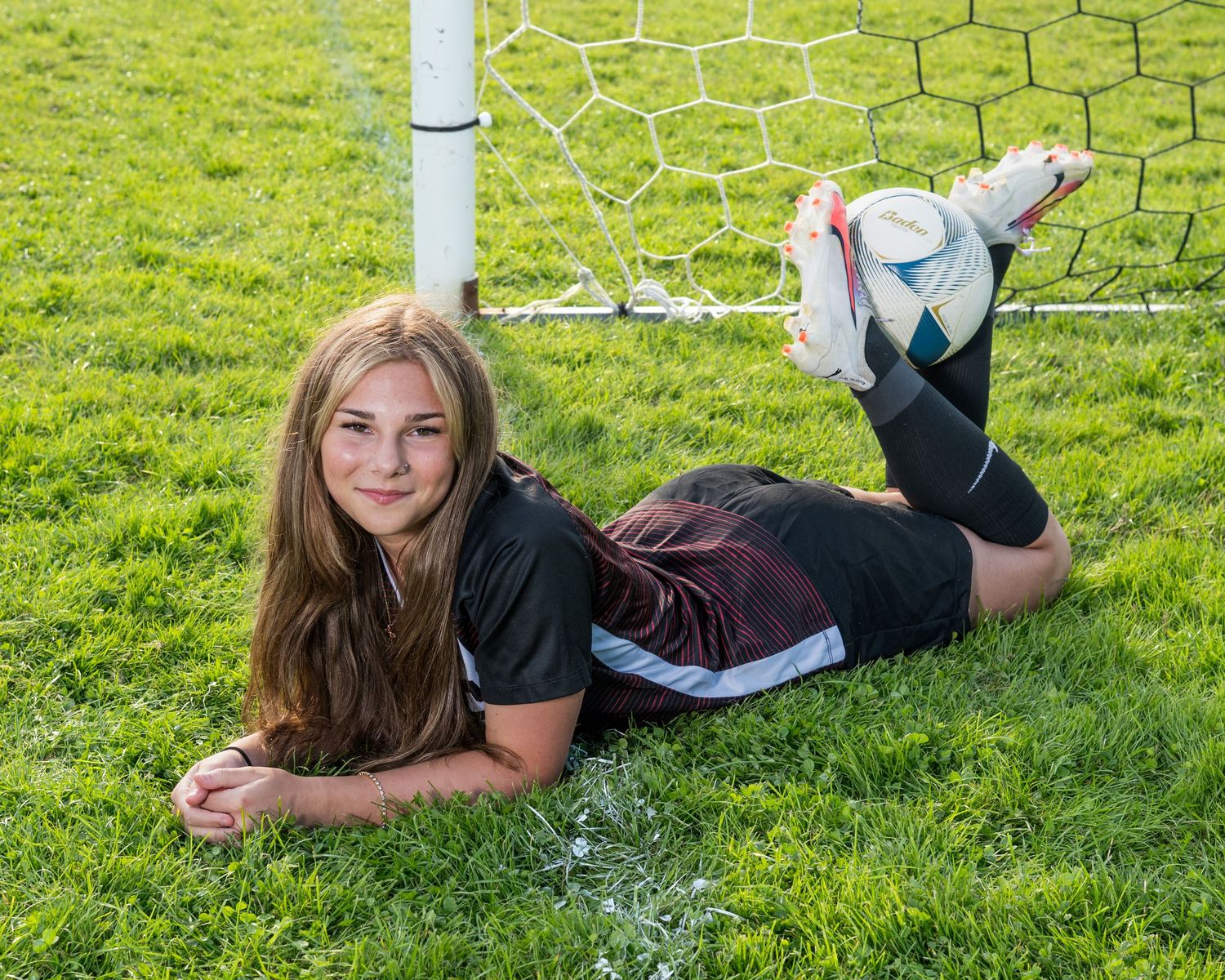 A student athlete poses on green grass near a chain link fence.