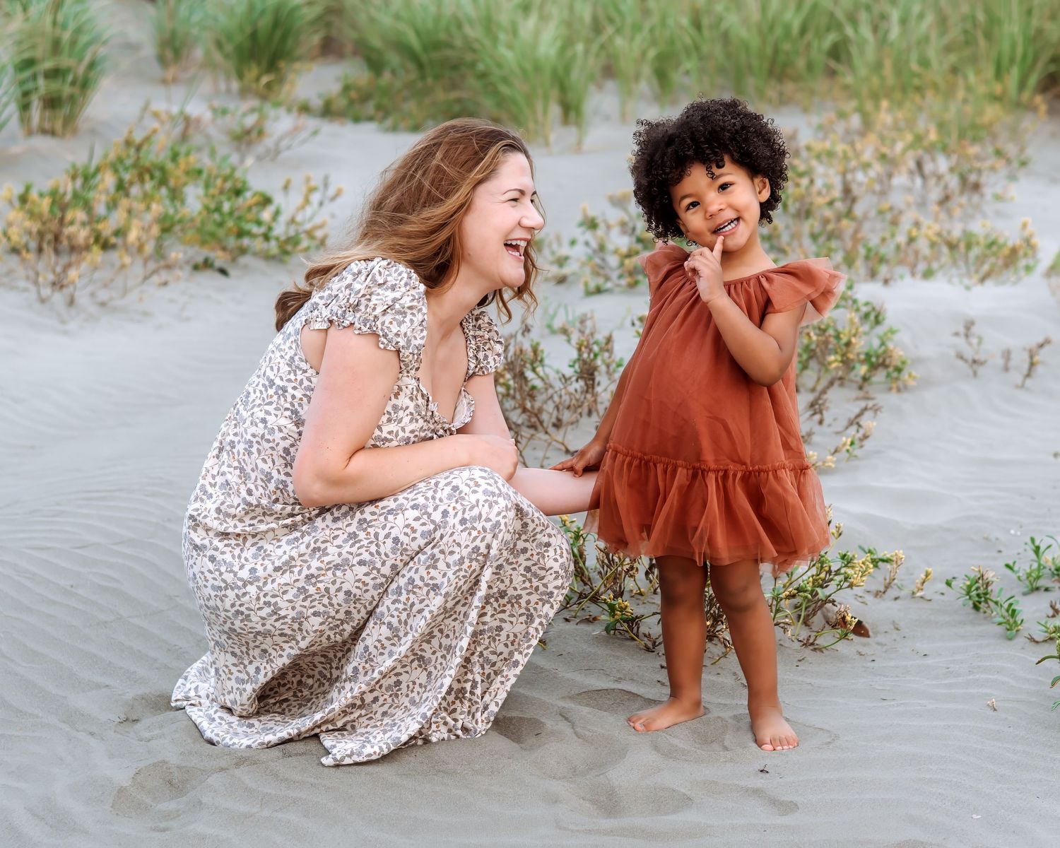 A laughing woman in a floral dress crouches beside a young girl in a rust dress on a sandy beach.