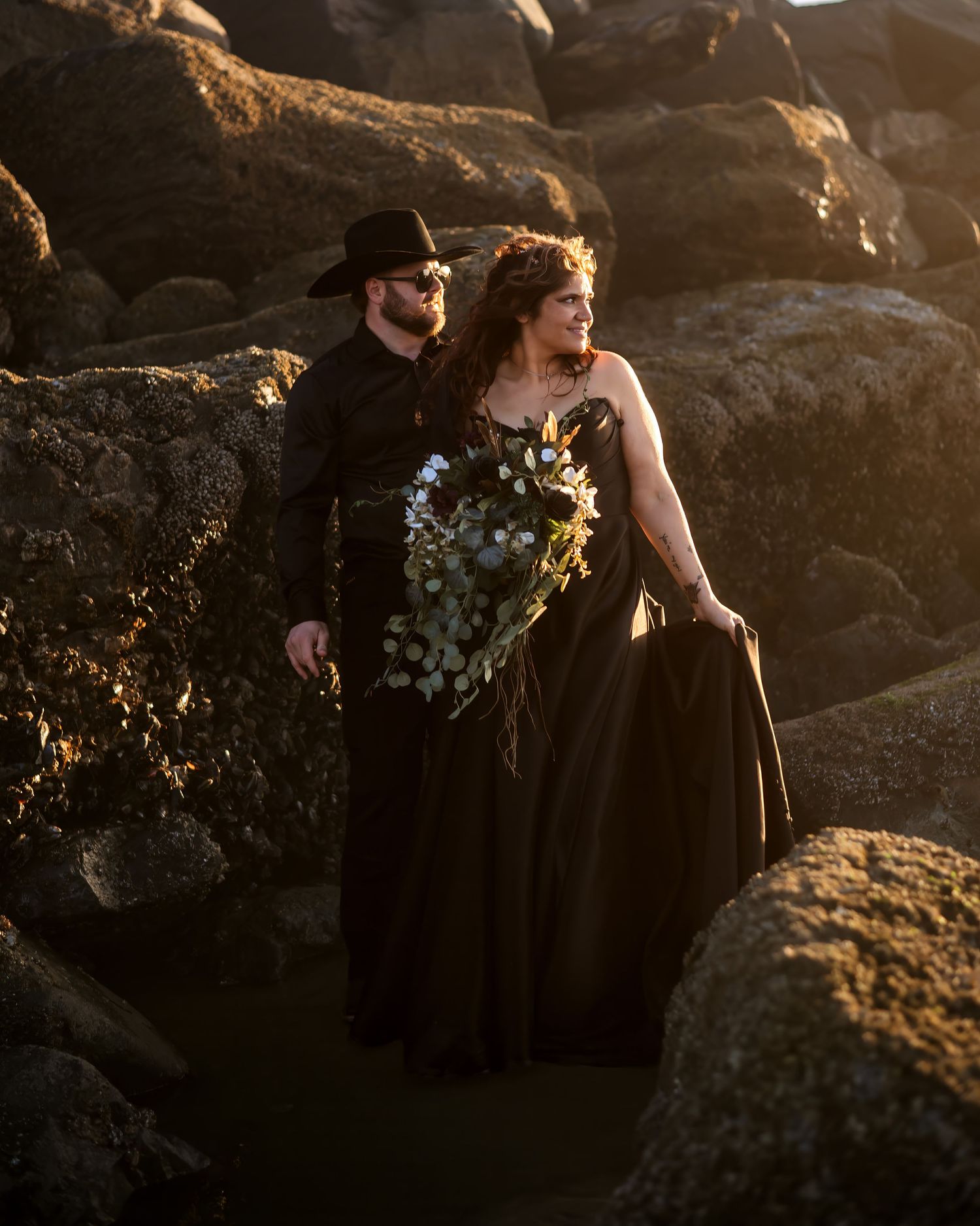 Bride in a black gown holding a greenery bouquet stands beside a groom in a black hat and shirt among mossy coastal rocks.