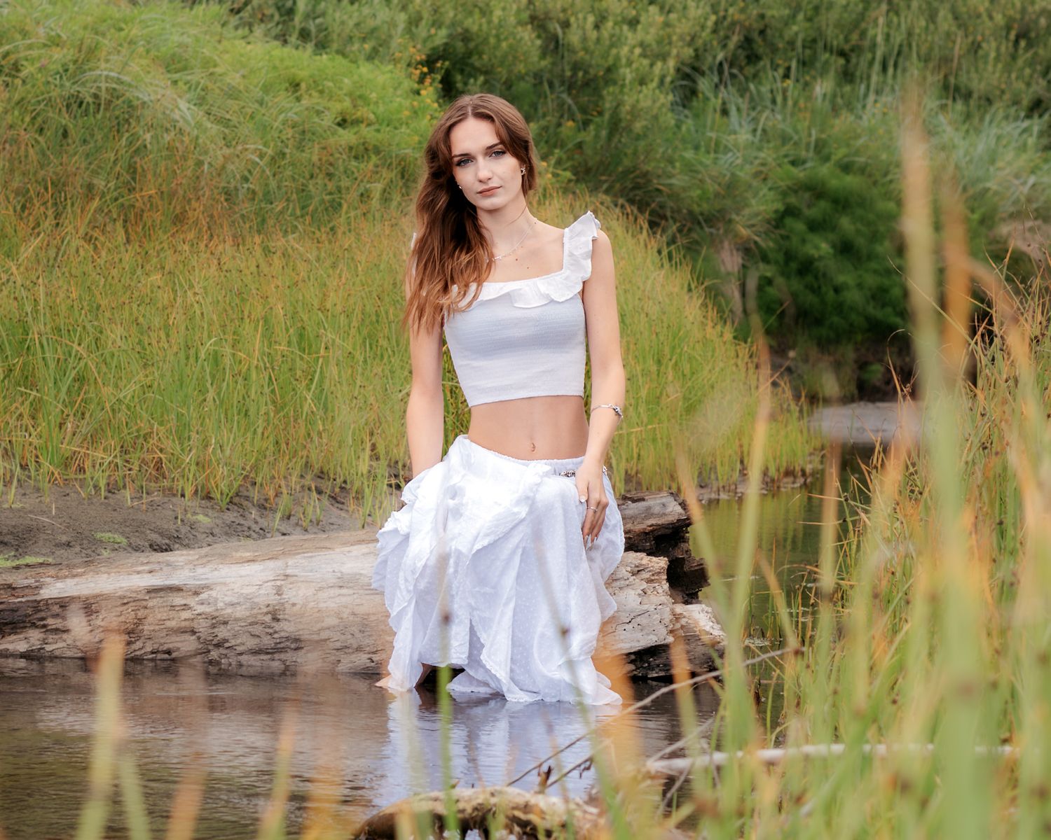Young woman in white crop top and flowing skirt sitting on driftwood by a serene river surrounded by lush greenery.
