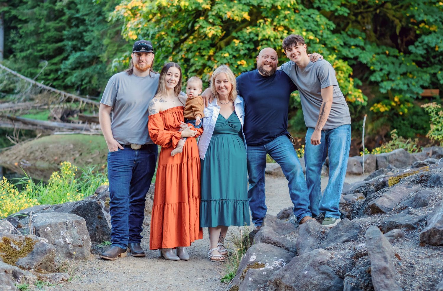 Family of six posing outdoors on rocky path surrounded by lush green trees in a Pacific Northwest forest setting.