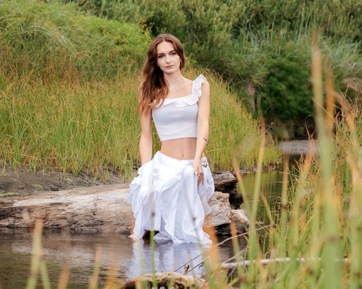 A woman in white clothing poses gracefully by a pond surrounded by tall grass.