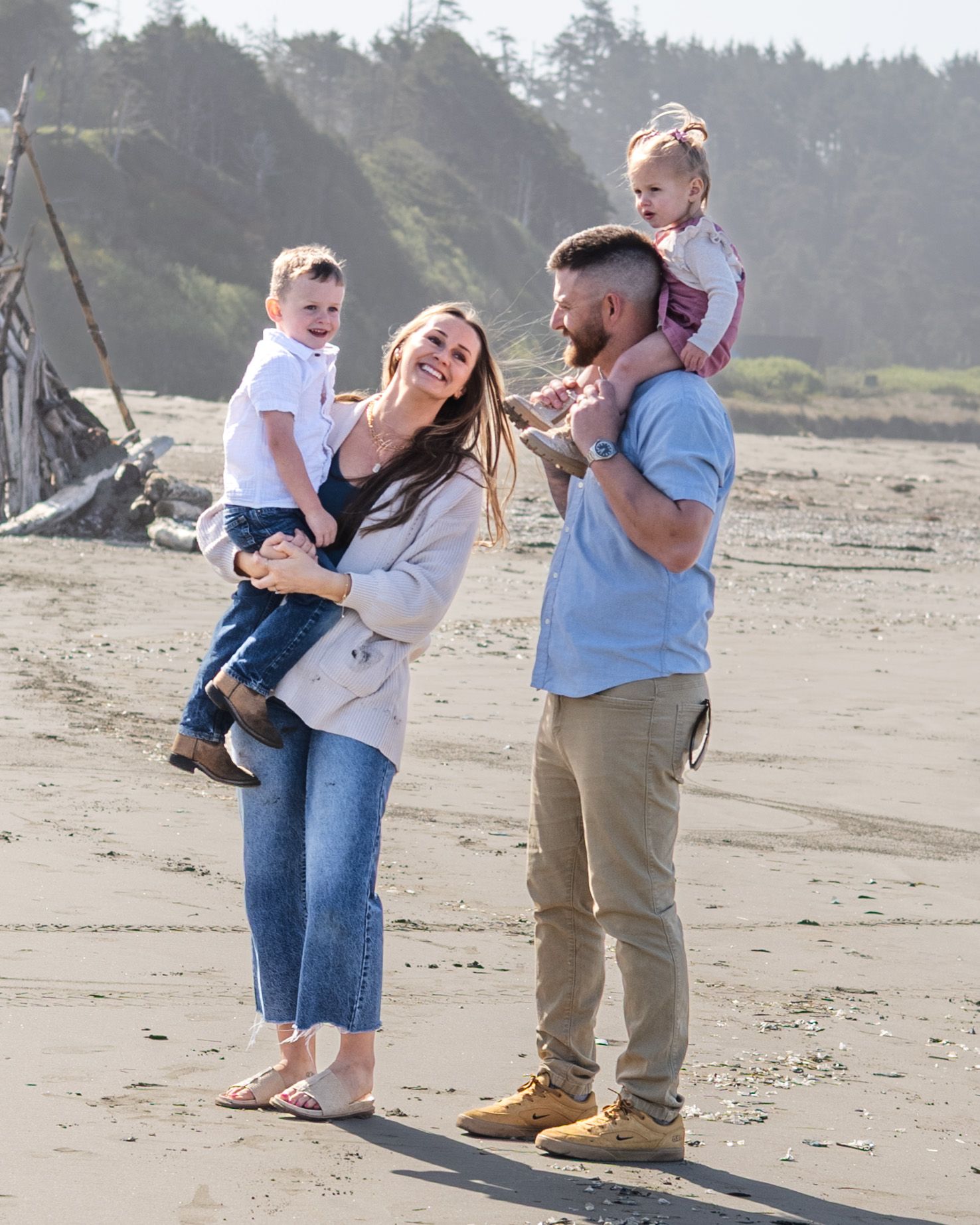Happy family of four enjoying a sunny beach day, parents holding two toddlers while smiling outdoors.