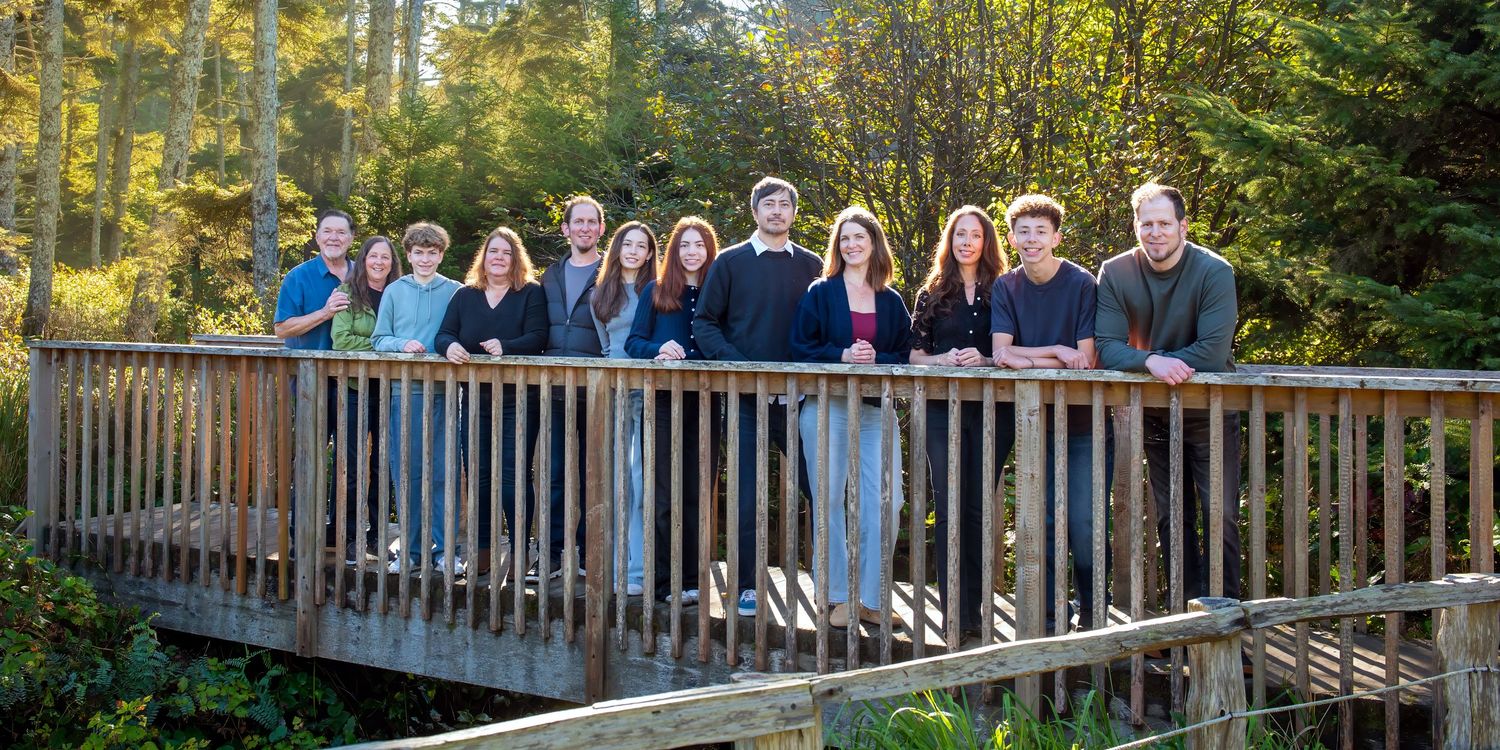 Group of twelve people standing side by side on a wooden footbridge surrounded by tall evergreen trees and greenery in a forest.