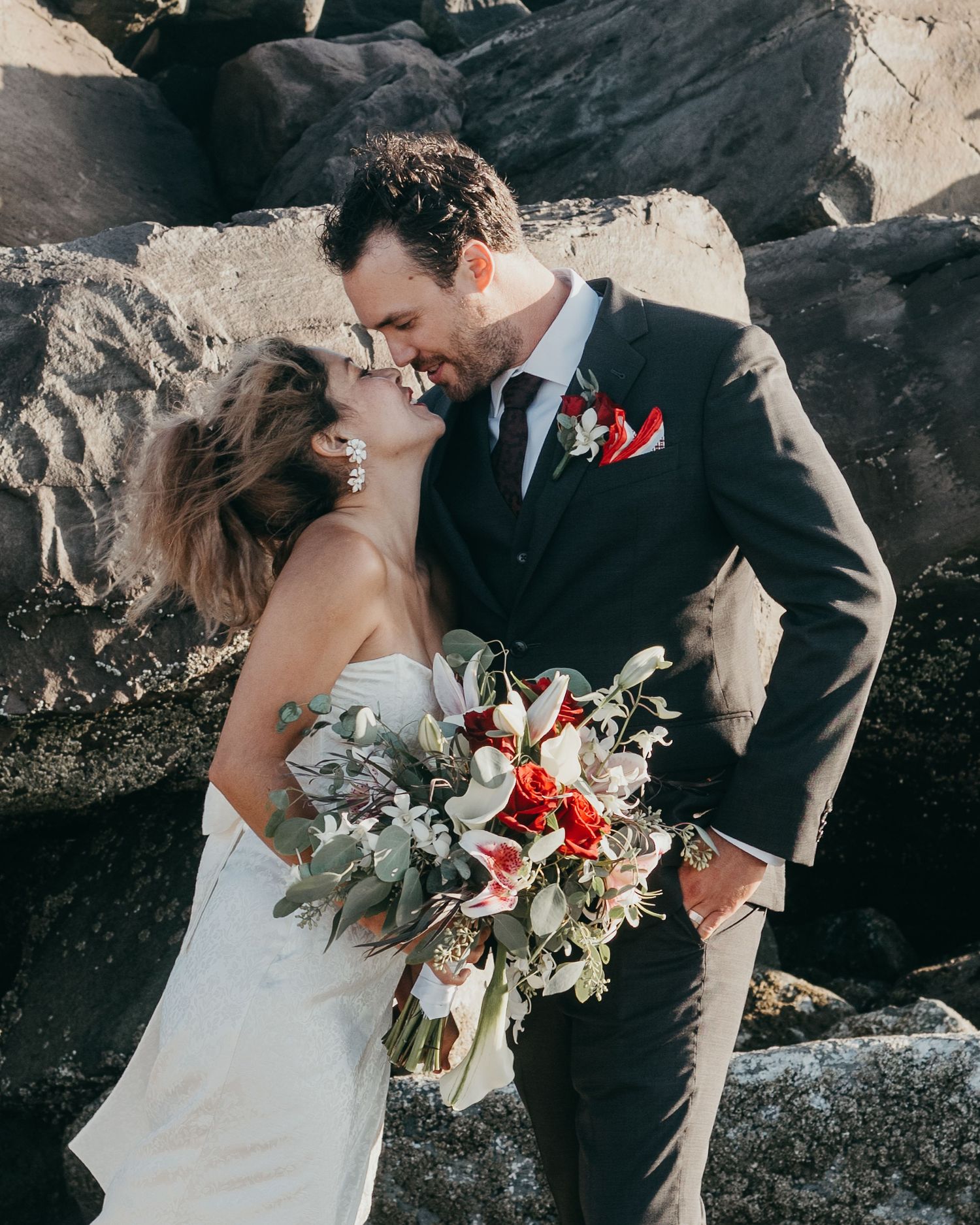 Romantic wedding couple poses against rocky backdrop with red and white floral bouquet.