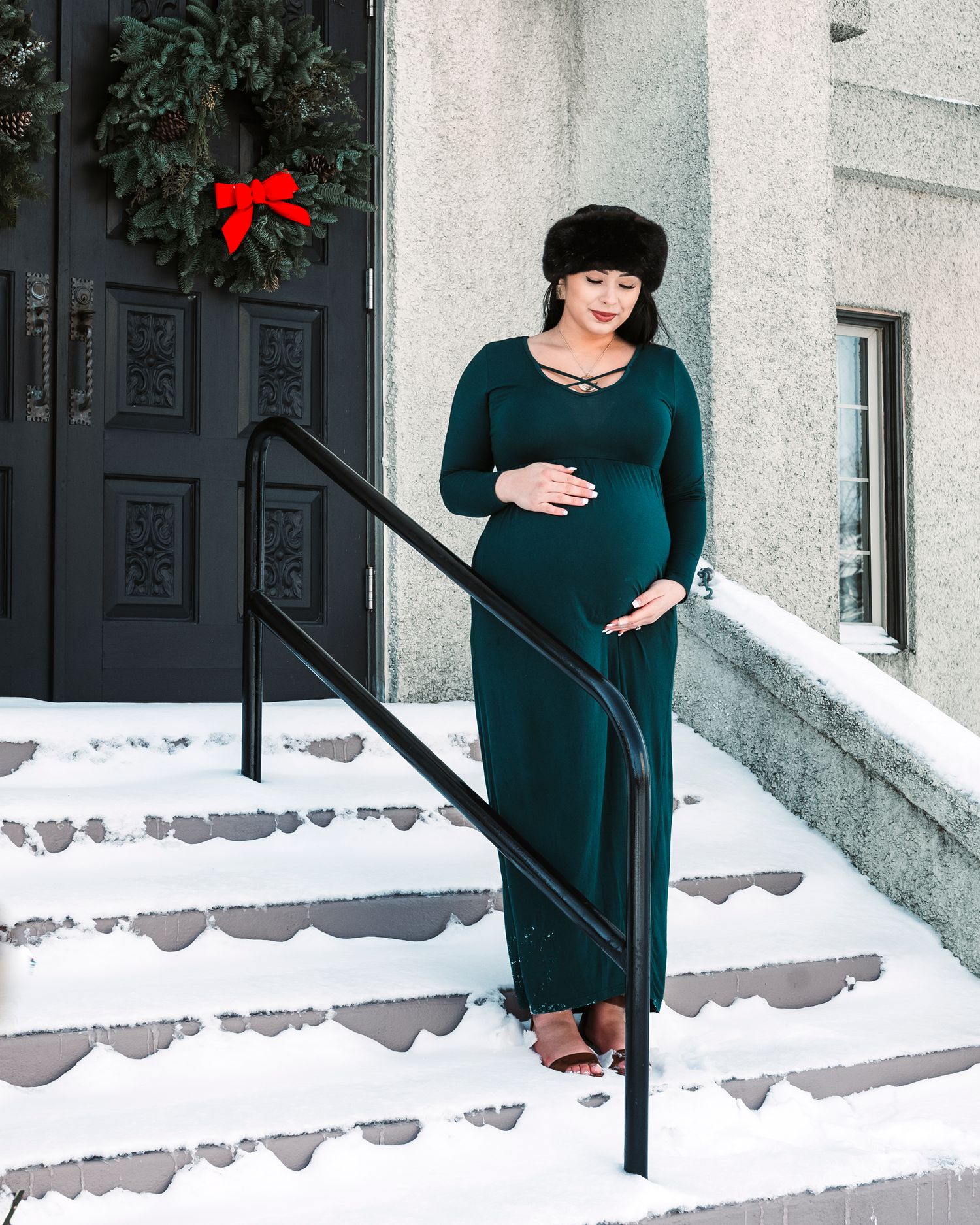 A pregnant woman in a dark green dress stands on snowy steps with a red wreath on the door behind her.
