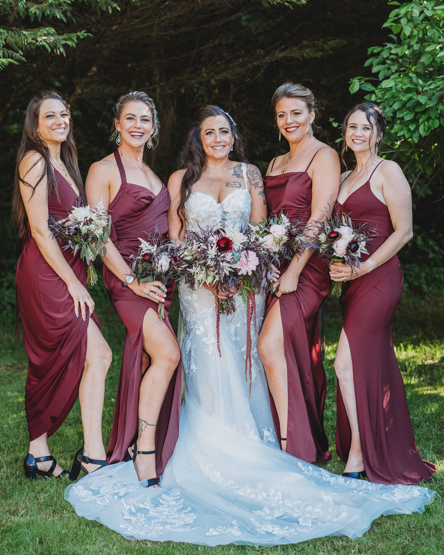 Bridesmaids in burgundy dresses pose with bride in white gown holding coordinated floral bouquets.