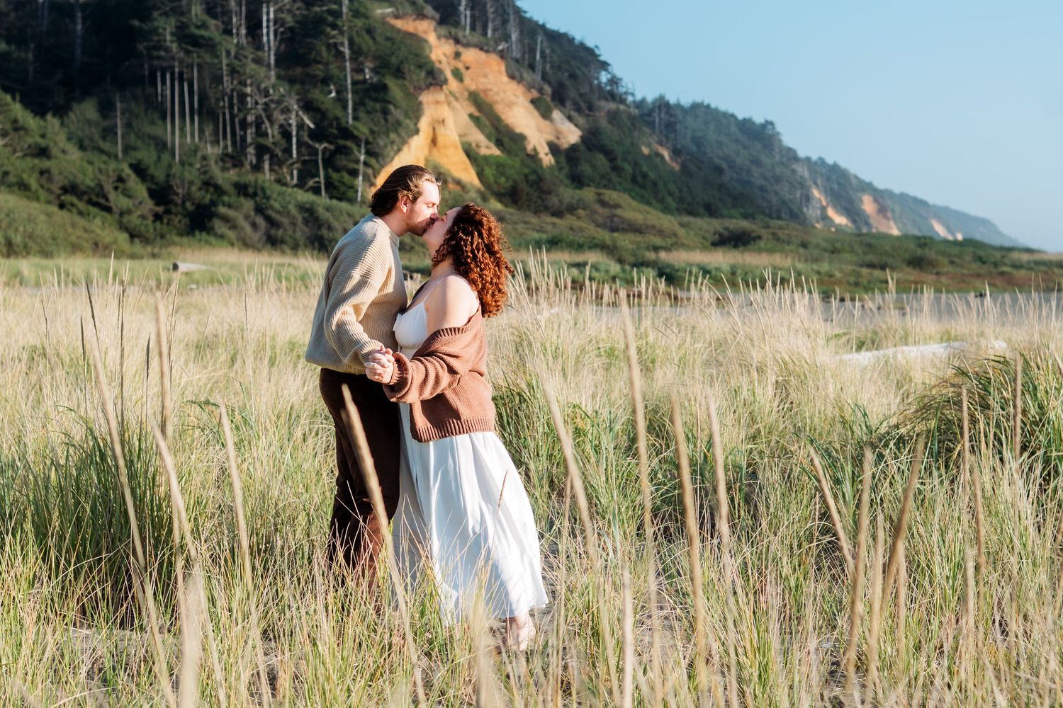 Couple embraces in scenic coastal field with mountains in background.