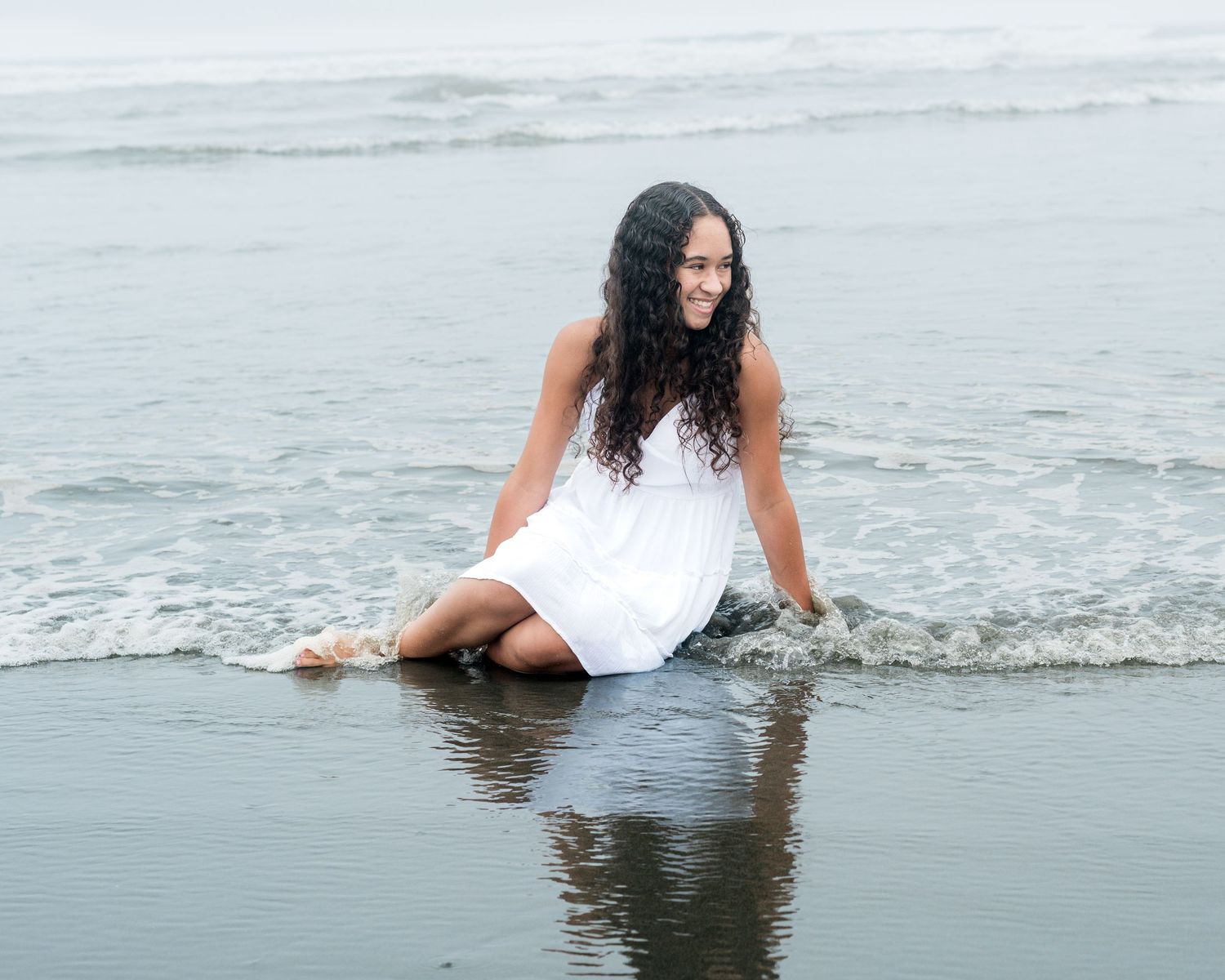 A woman in white dress sits at the edge of ocean waves on a beach.