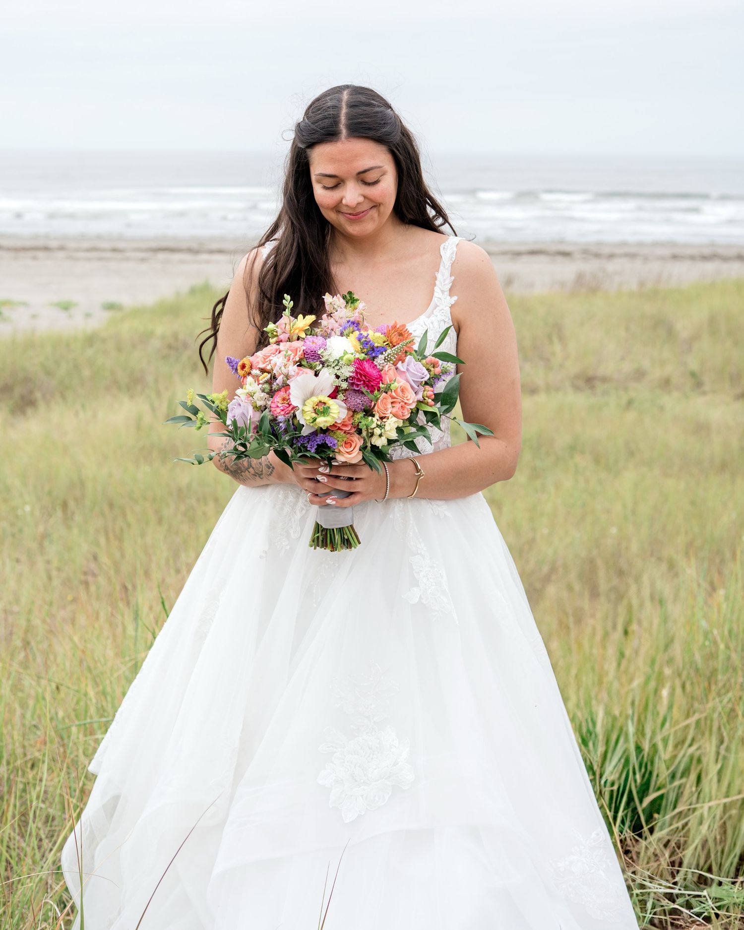 Bride holding vibrant floral bouquet poses in white wedding dress against coastal landscape.