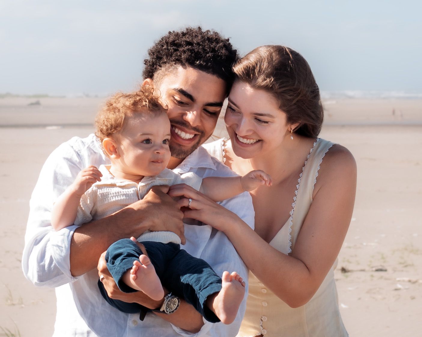 Happy young family with baby smiling together on a sunny beach, parents lovingly holding their toddler.