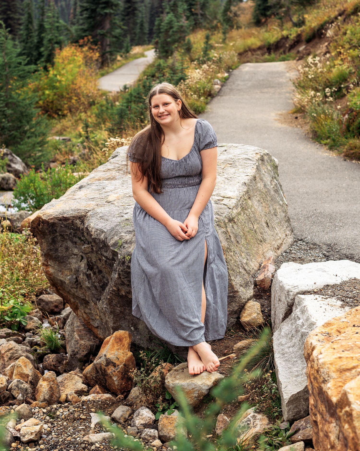Person in gray dress sitting on large rocks with mountain trail and evergreen forest backdrop.