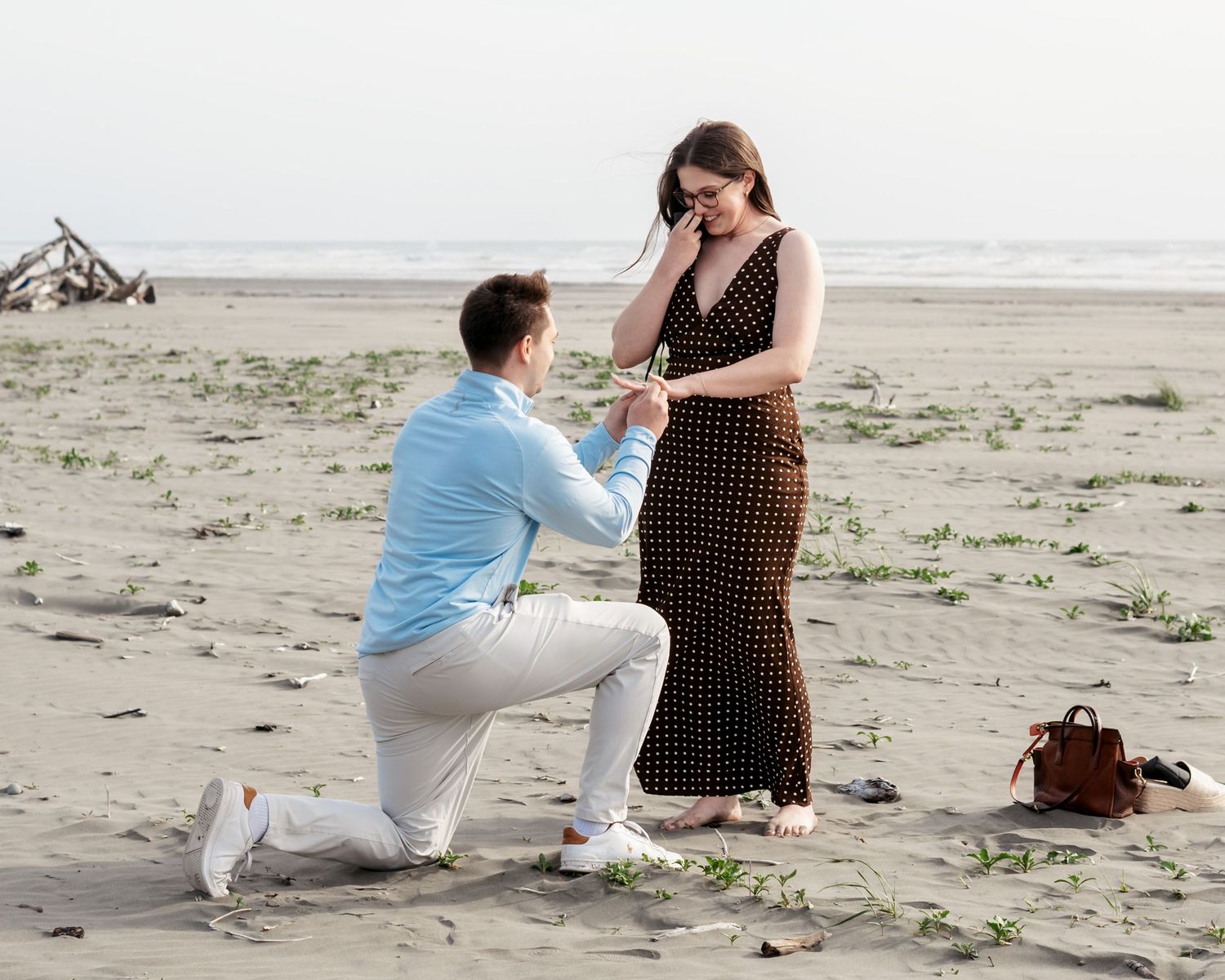 Man kneeling on beach proposing to woman in polka dot dress, romantic marriage proposal on sandy shore.