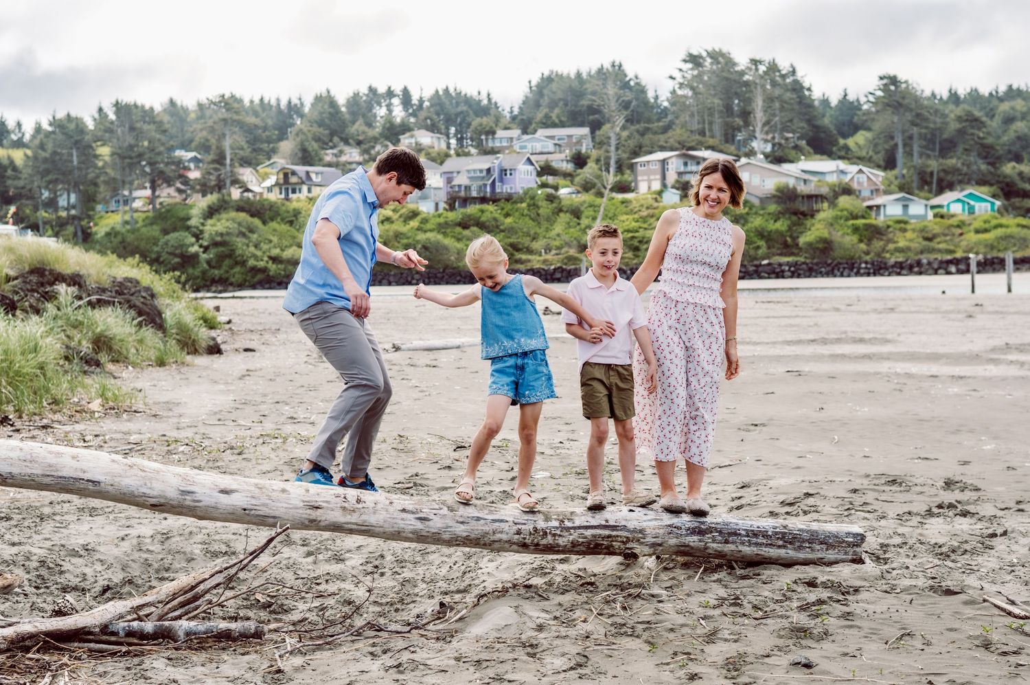 Family of four playing on driftwood at a sandy beach with lush green trees and coastal homes in the background.