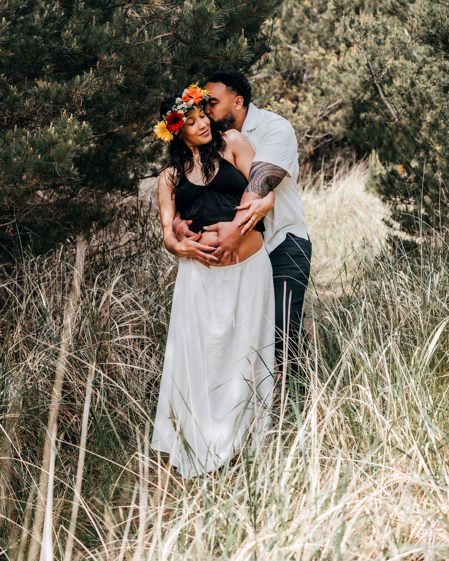 Couple embracing in tall grass, pregnant woman wearing flower crown and white skirt, partner kissing her head outdoors.