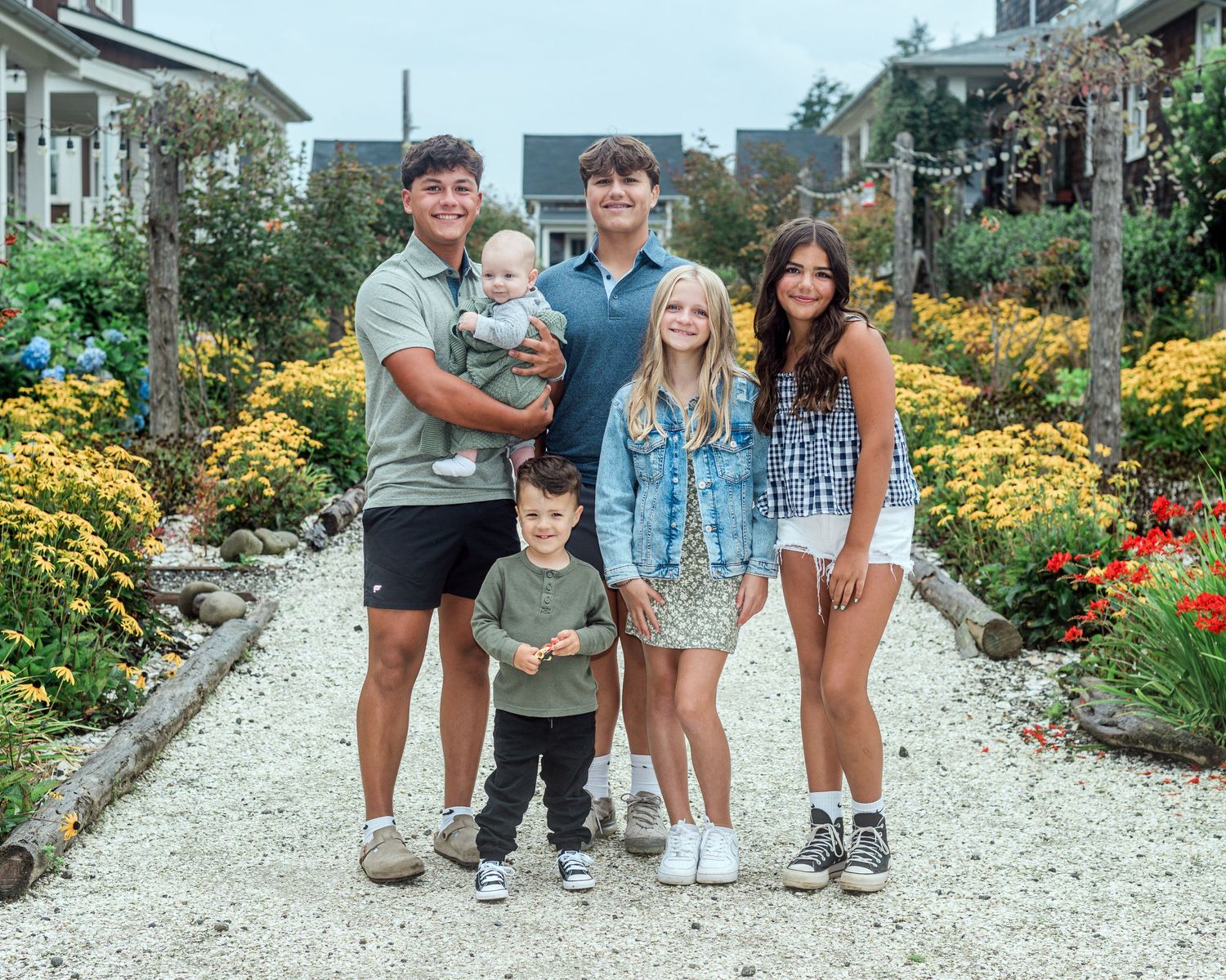 Family of six posing on a garden path surrounded by colorful flowers on a sunny day.