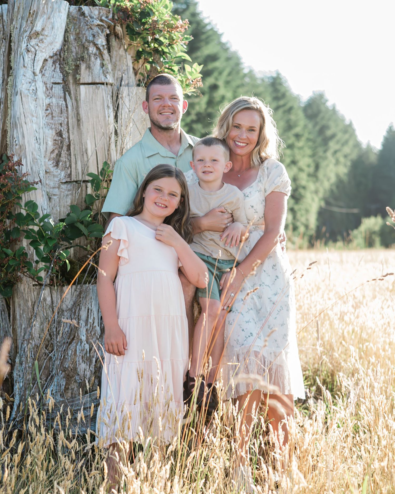 Family of four smiling outdoors near a rustic tree in a golden field with evergreen trees in the background.