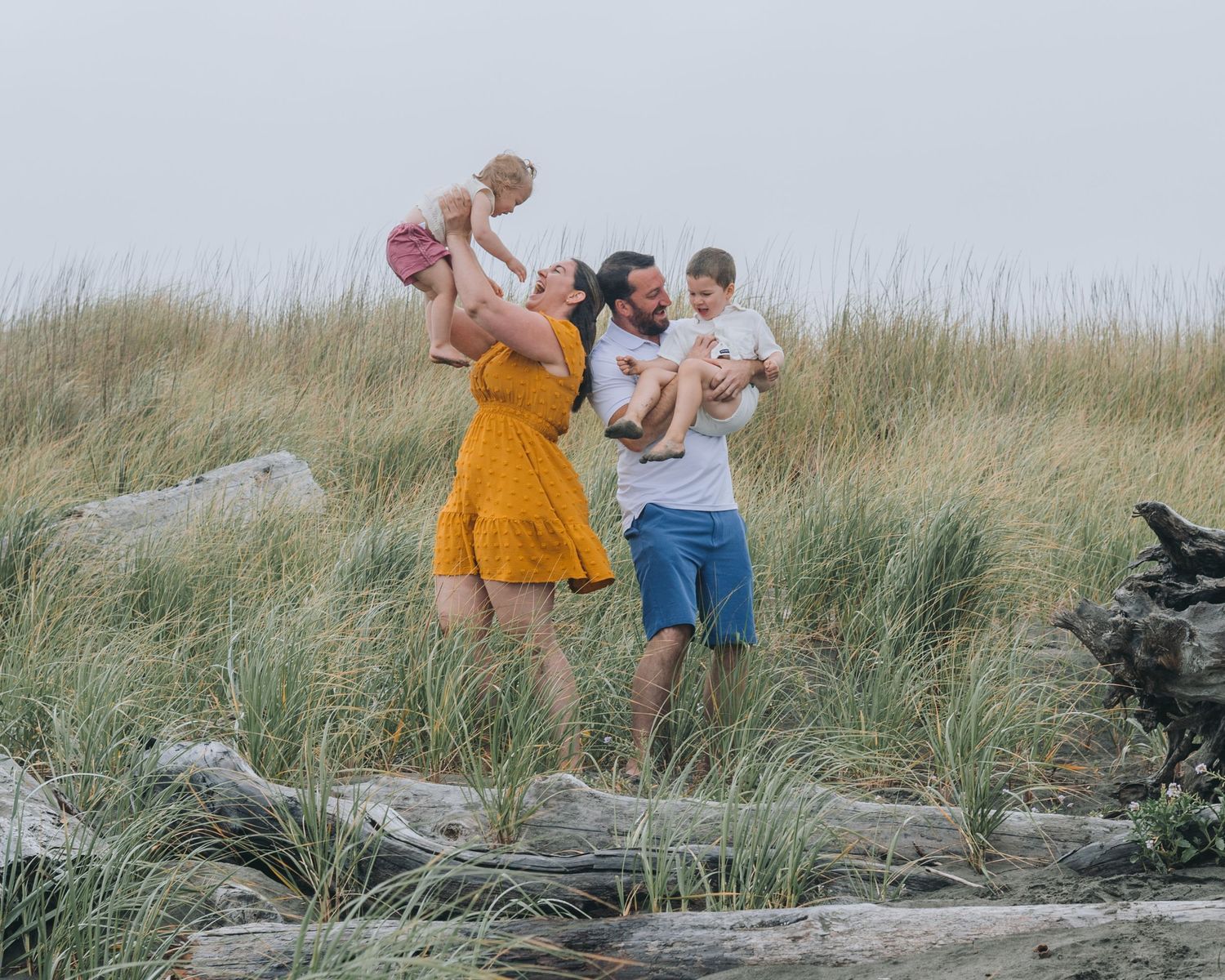 A couple and their two children spendinf together on a beach path surrounded by tall grass and driftwood at sunset.