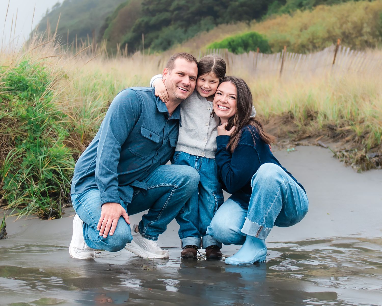 Family of three smiling and crouching in shallow water on a beach, wearing casual blue outfits on a cloudy day.