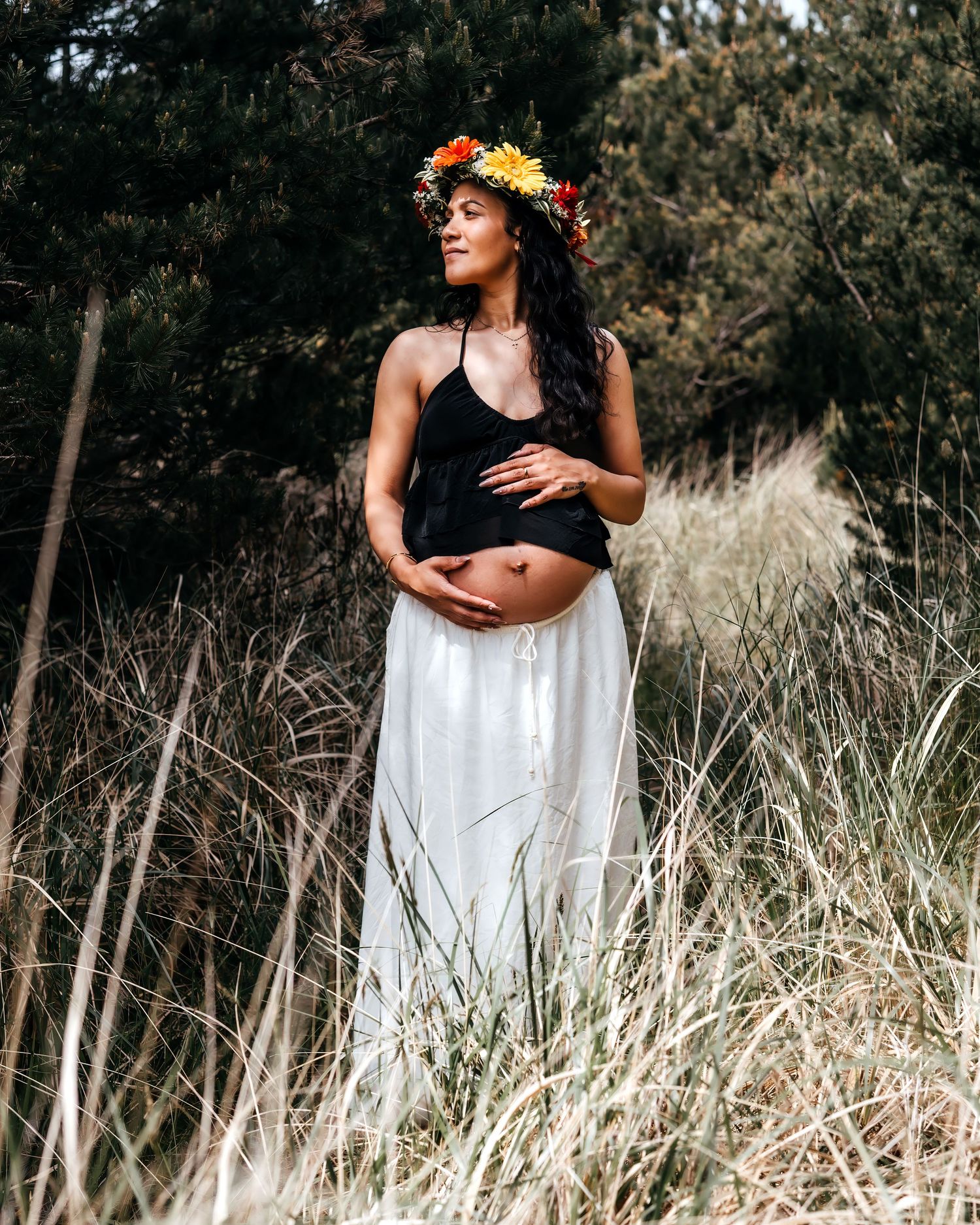 Pregnant woman in black crop top and white skirt wearing flower crown standing in tall grass outdoors.