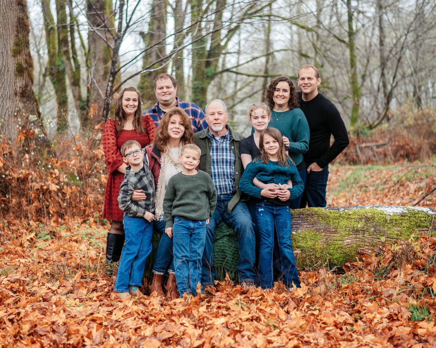 Family portrait of multi-generational group posing outdoors among autumn leaves and bare trees in a forest setting.