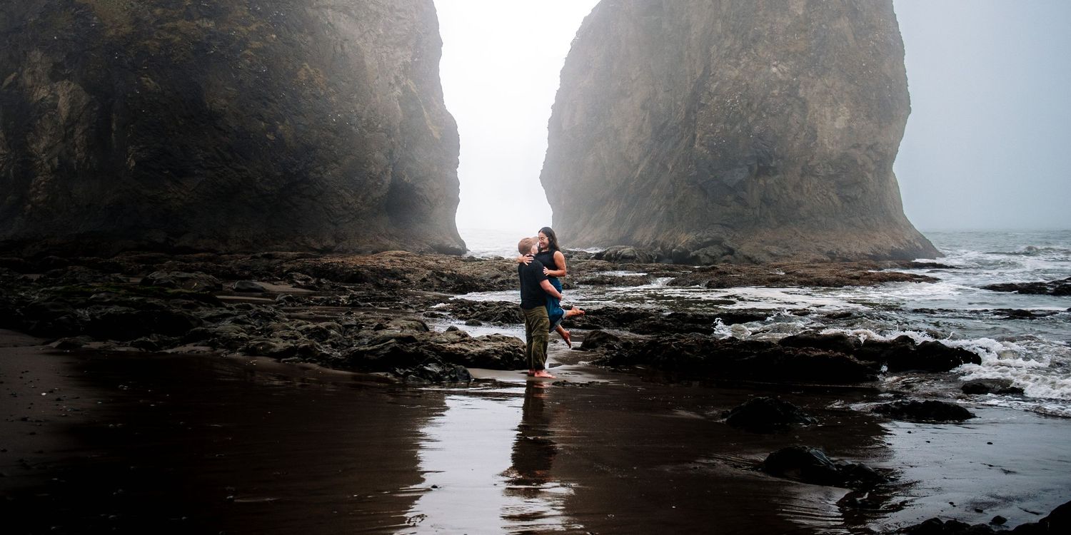 Person standing on a rocky beach with dramatic sea stacks rising through misty fog on the Oregon coast.
