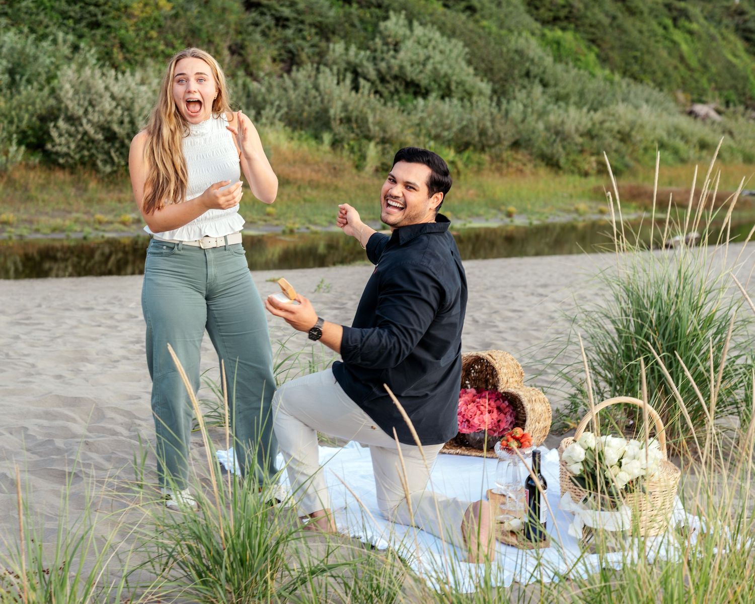 A surprise proposal moment captured on a scenic beach with picnic setup.