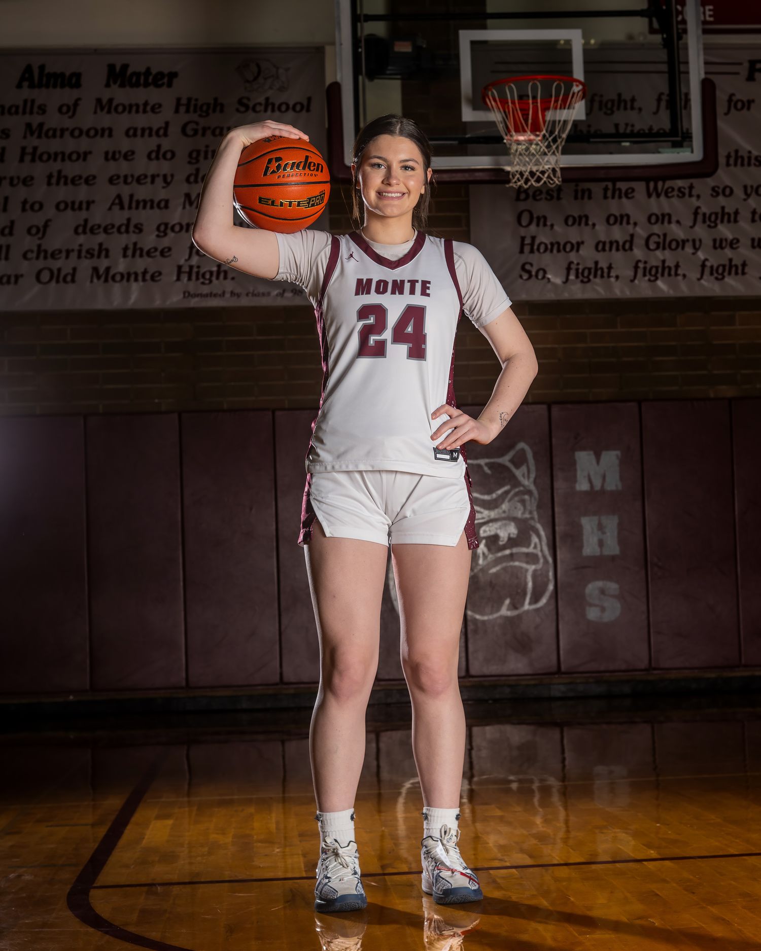 Basketball player poses in white uniform on dark gymnasium court with dramatic lighting.