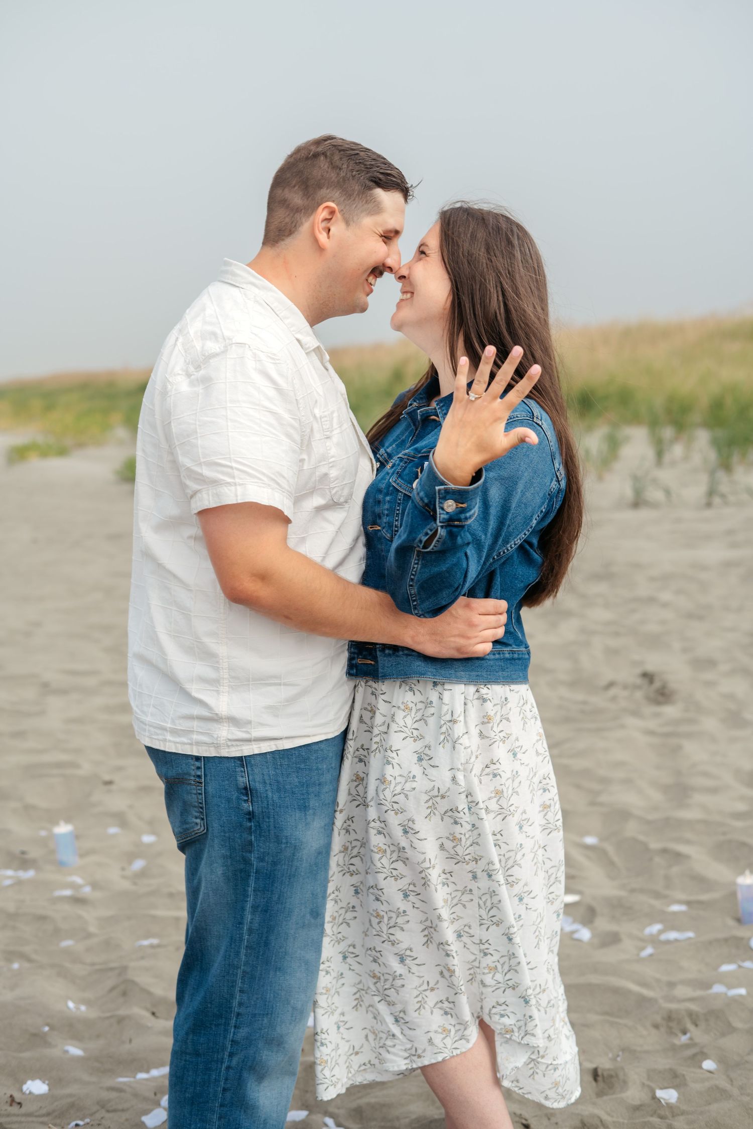 A couple embraces on a beach while showing off an engagement ring.