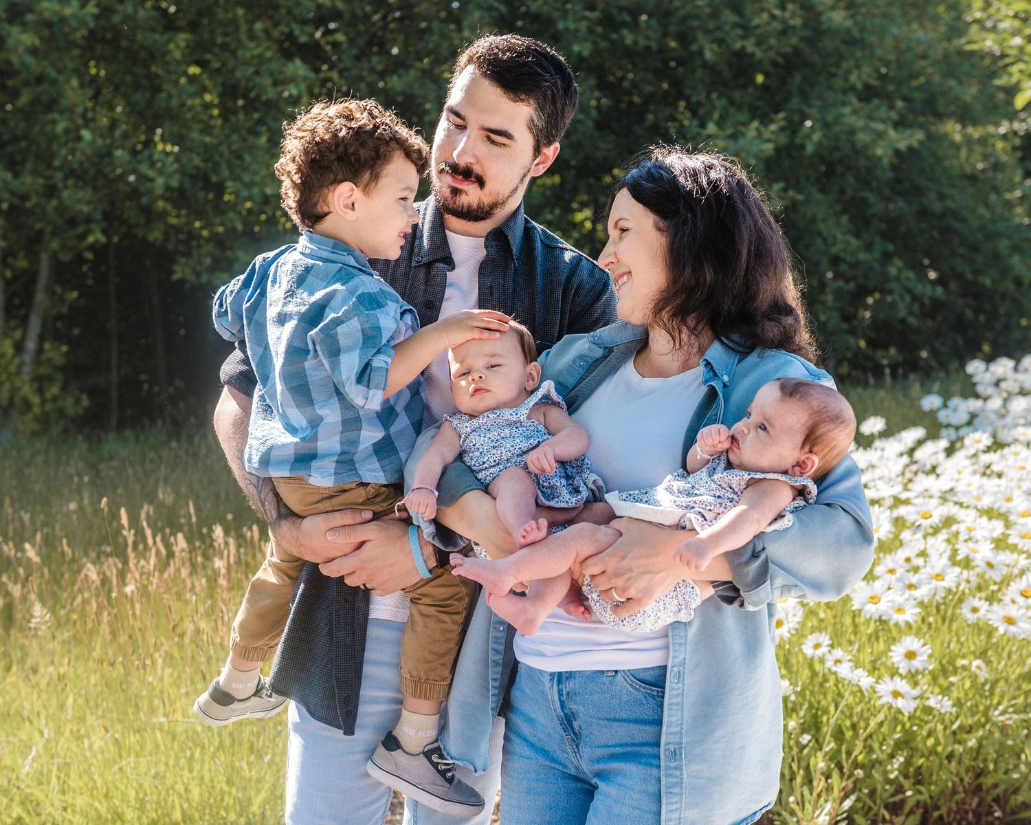 Happy young family with toddler and twin babies posing outdoors in a sunny meadow with wildflowers and green trees.