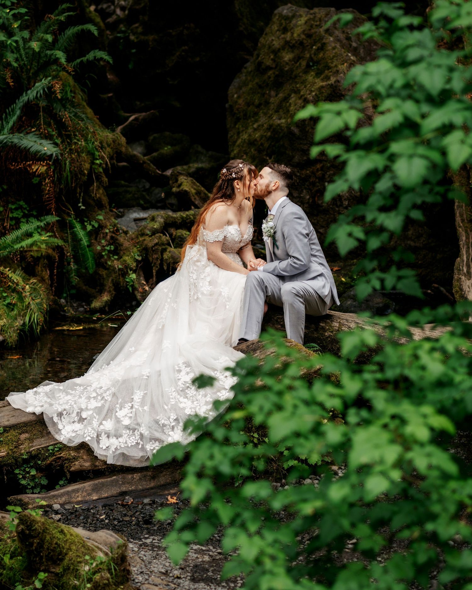 Bride in a lace wedding gown and groom in a gray suit kiss on a mossy log beside a stream in a lush green forest.