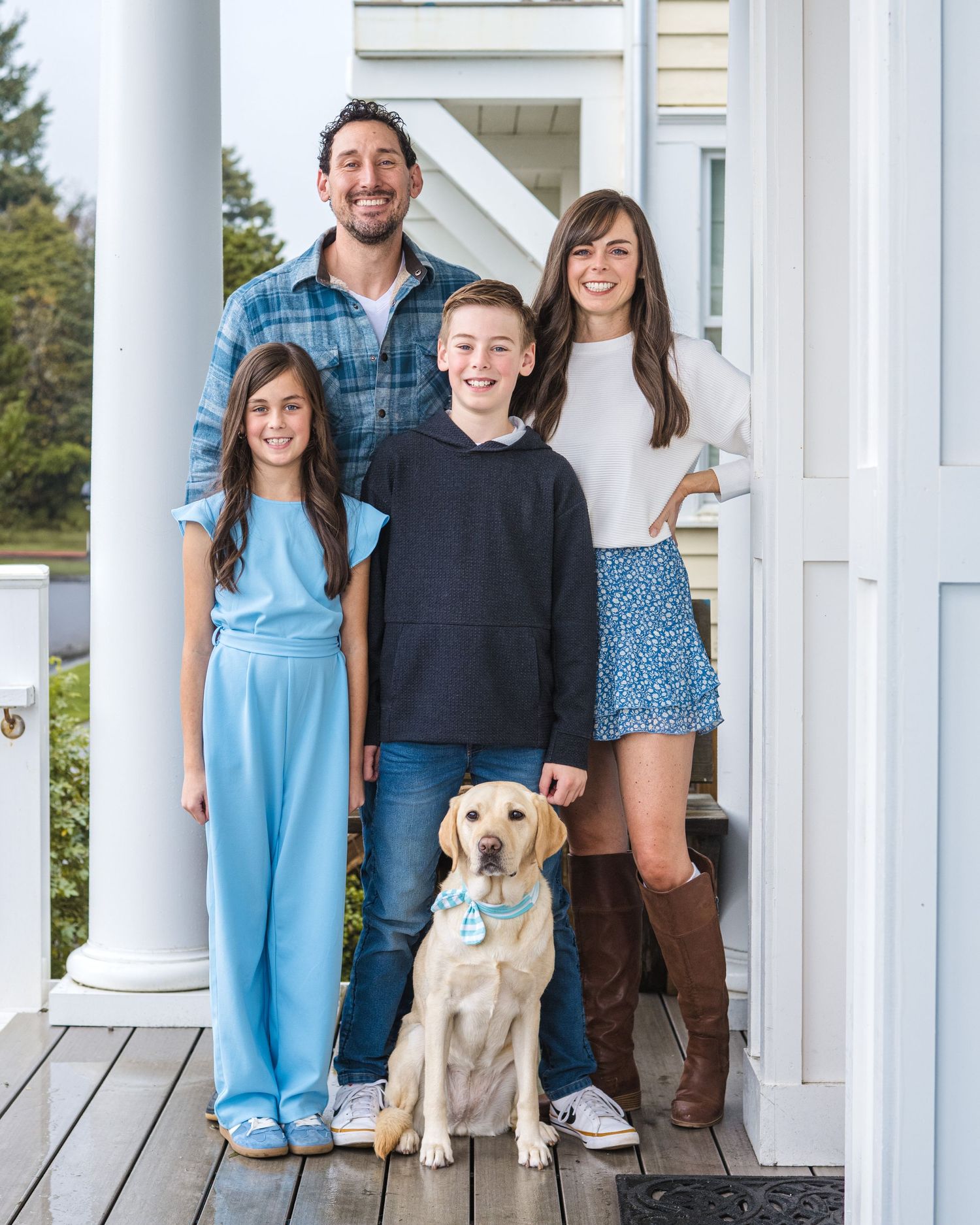 Family of four posing with a yellow Labrador on a white porch, smiling together in casual outfits.