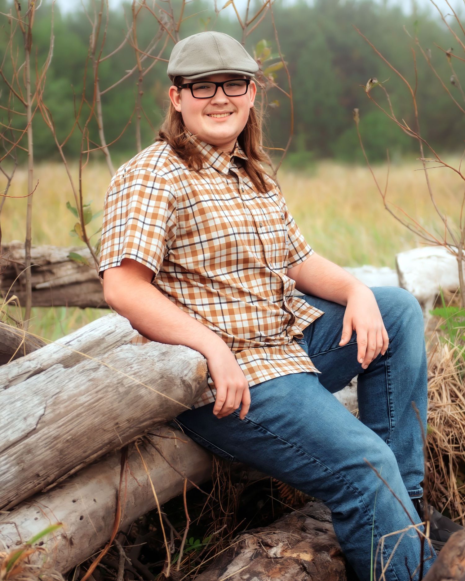 Smiling teen in a flat cap, glasses, plaid short-sleeve shirt and jeans leaning on a weathered log pile beside a grassy field and trees.