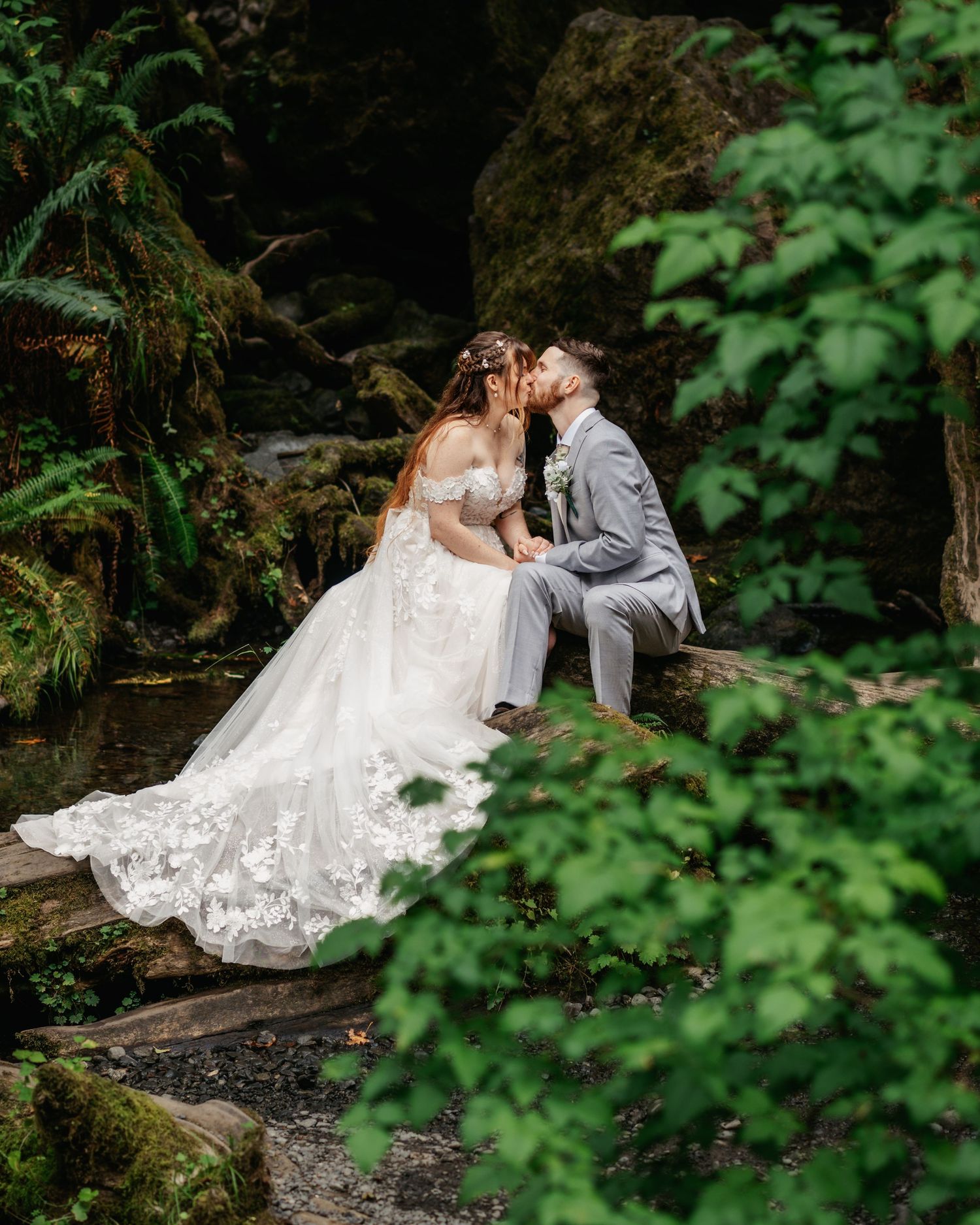 Bride and groom share a romantic kiss on mossy rocks in a lush forest setting during their wedding day.