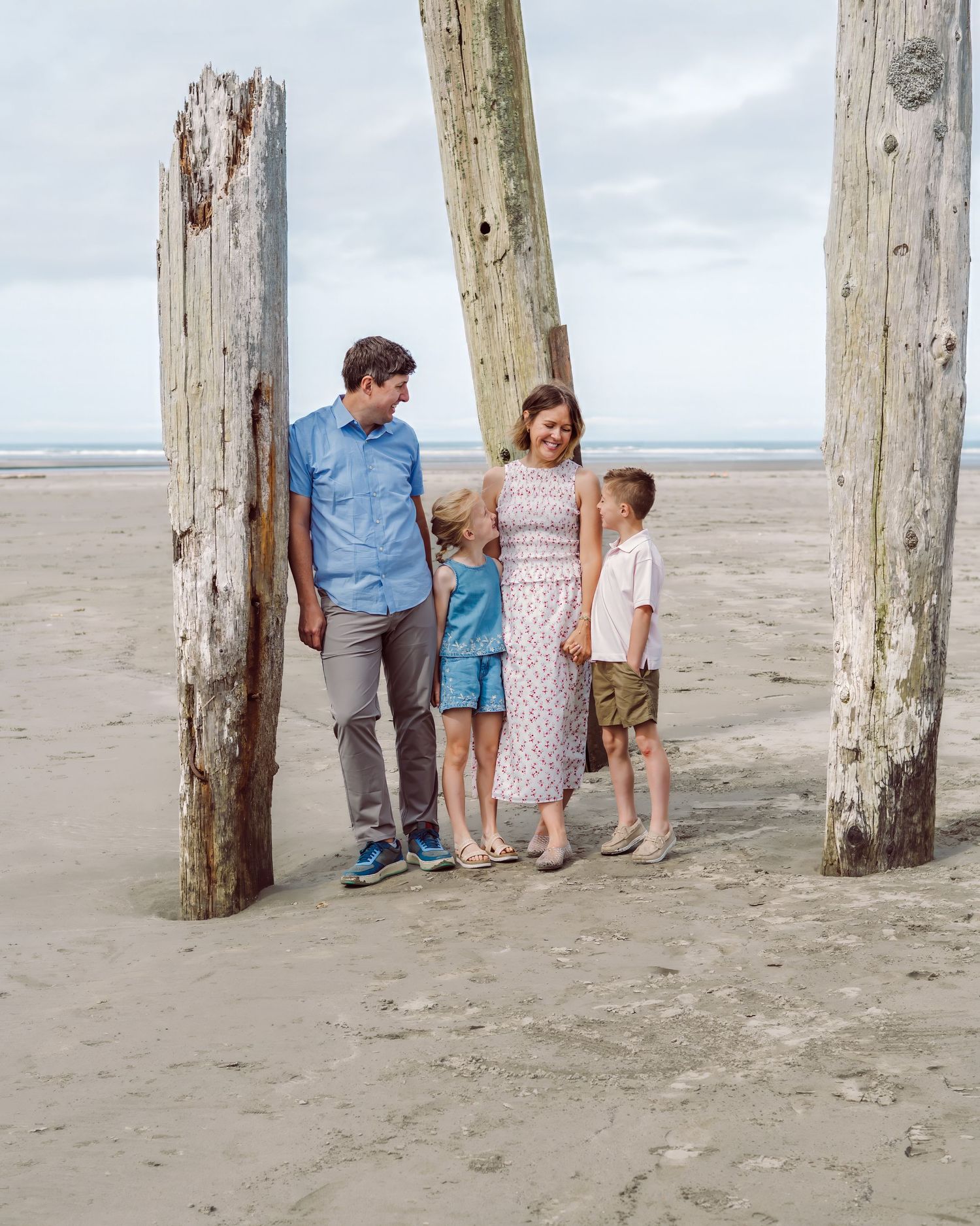 Family of four posing between weathered wooden posts on a sandy beach under a cloudy sky.