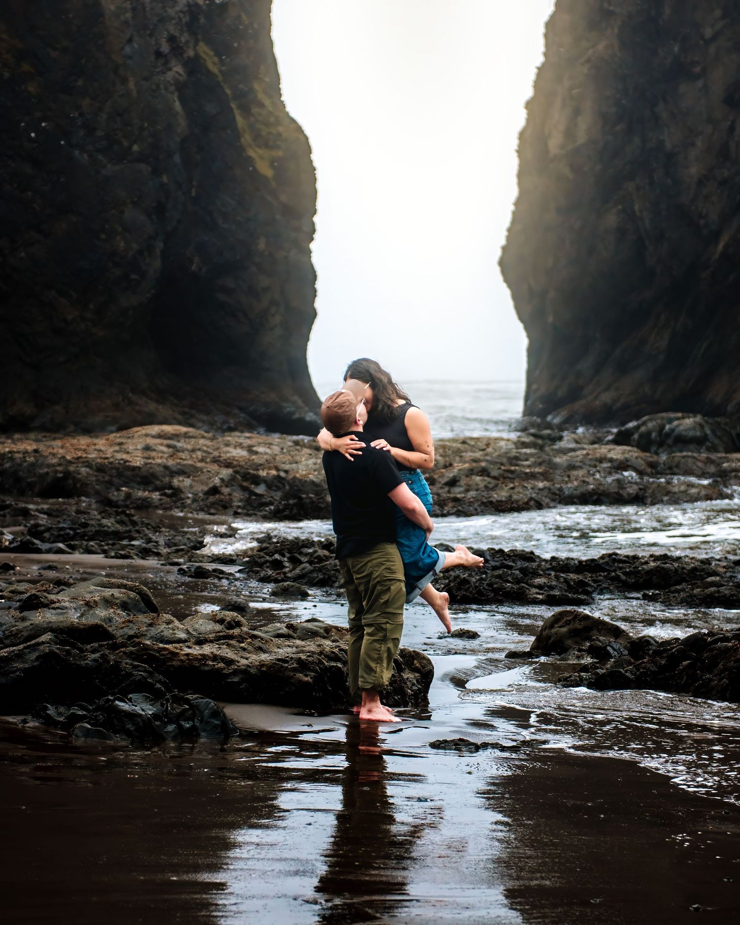 A couple embraces romantically between rocky sea cliffs on a misty beach, the man lifting his partner near tidal pools.
