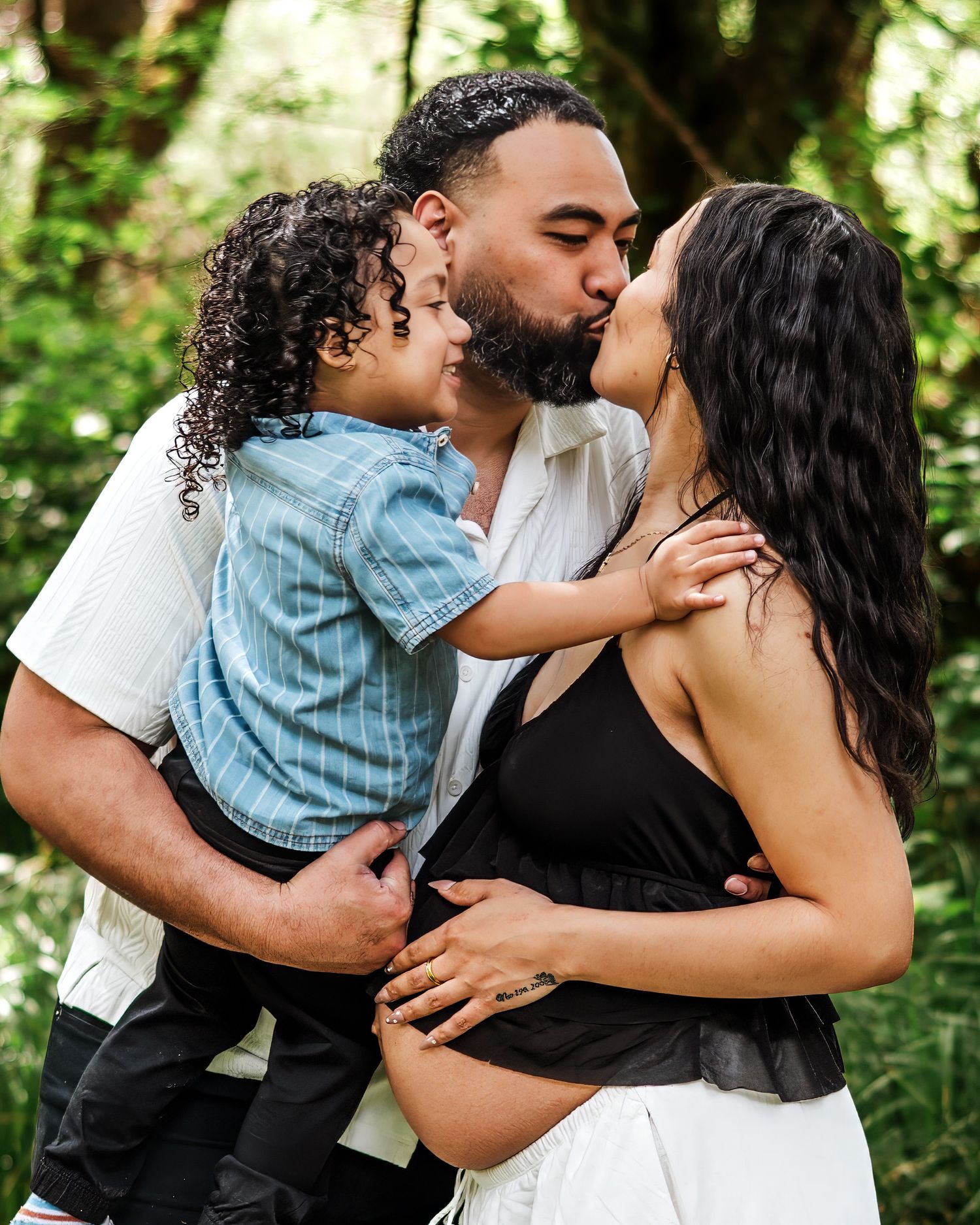 Couple kissing outdoors in lush green forest while man holds young curly-haired child, pregnant woman in black top.