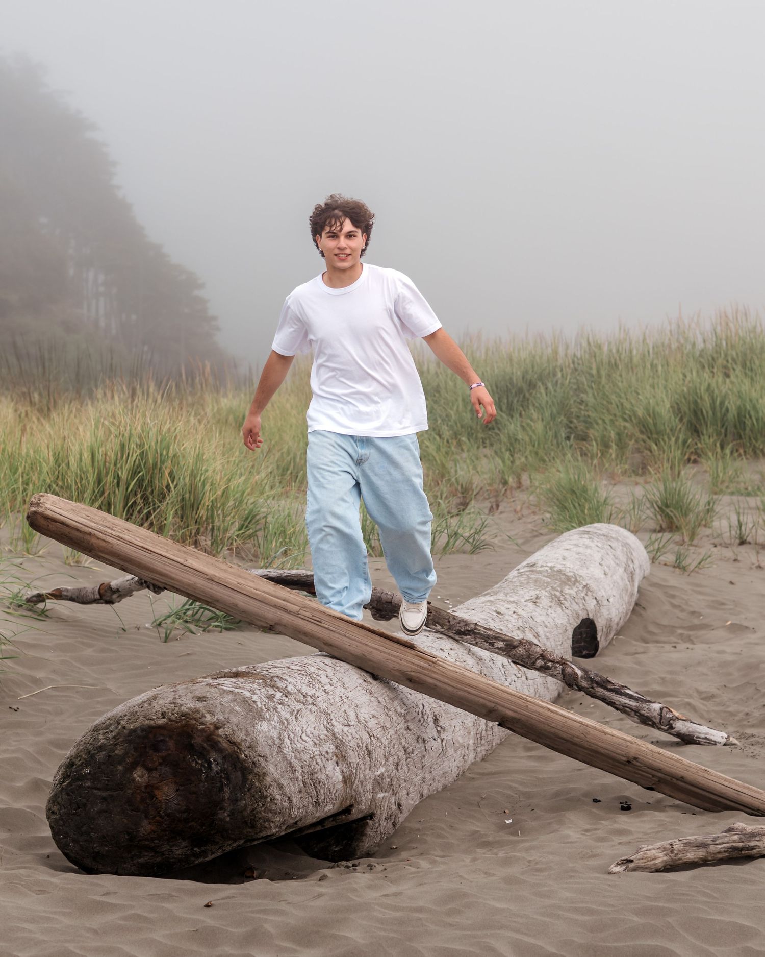 Person in white shirt and light pants walking on a fallen log at a misty beach.