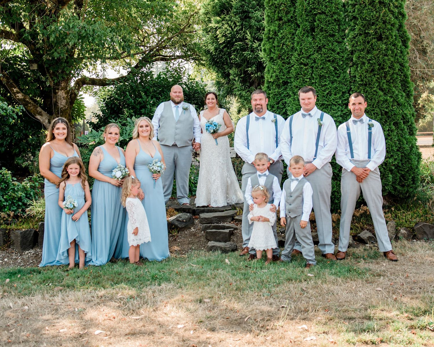 Wedding party in light blue and gray attire poses together against an ivy-covered wall.