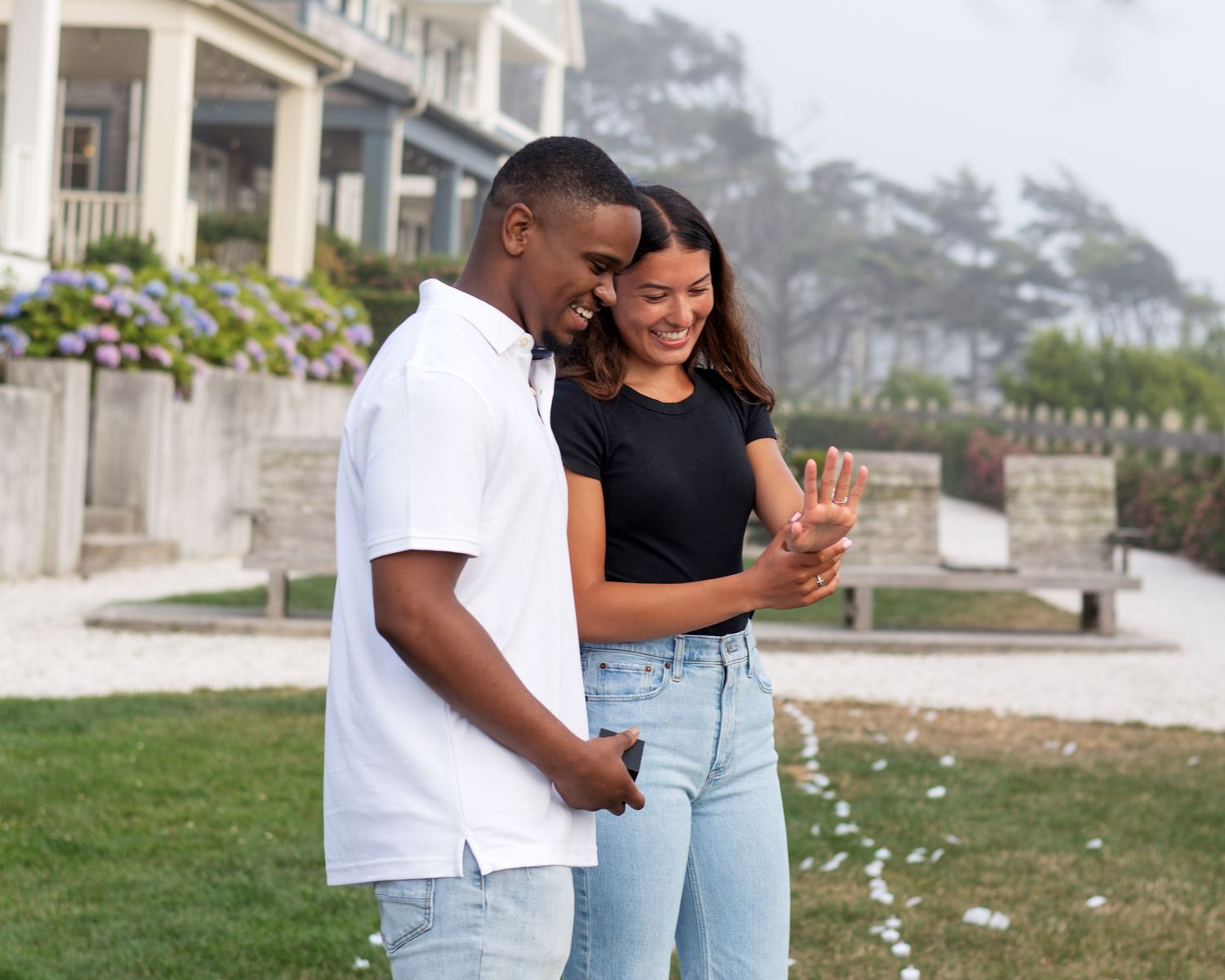 A couple embraces on a grassy lawn in front of elegant white buildings.