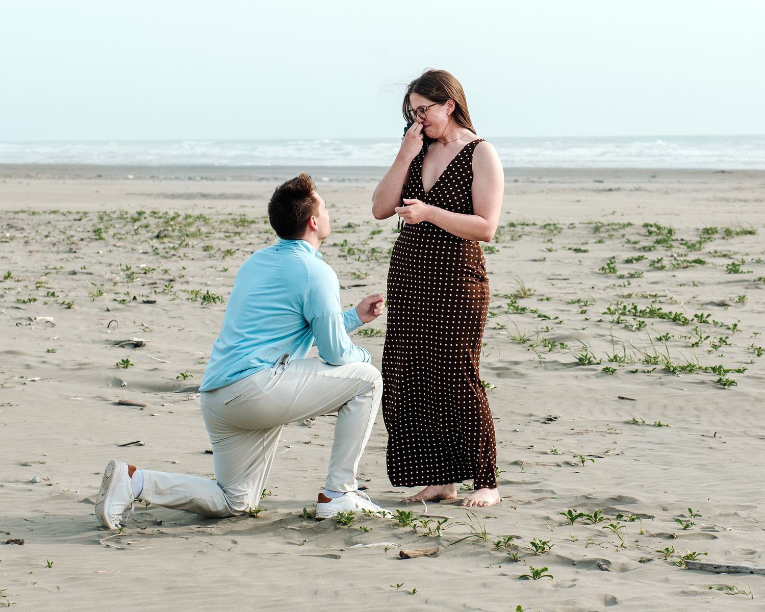 Man kneeling on beach proposing to emotional woman in polka dot dress by the ocean shore.