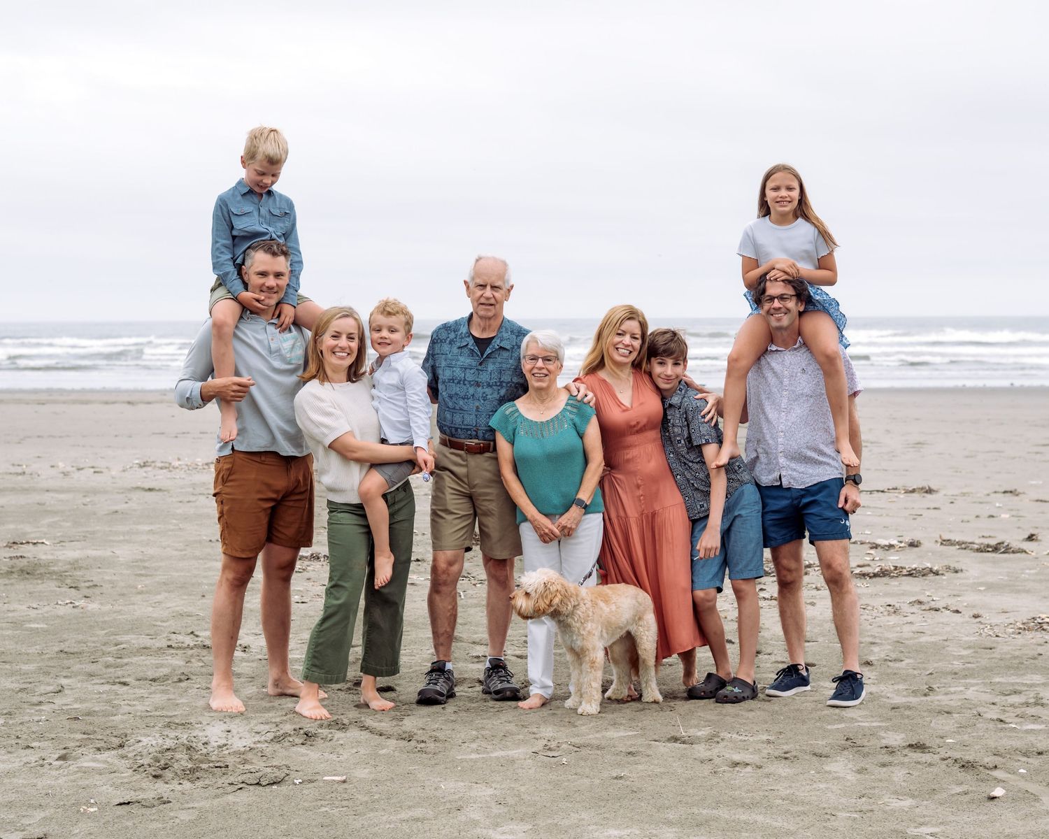 Large multigenerational family posing together on a sandy beach with a white dog, ocean waves in the background.