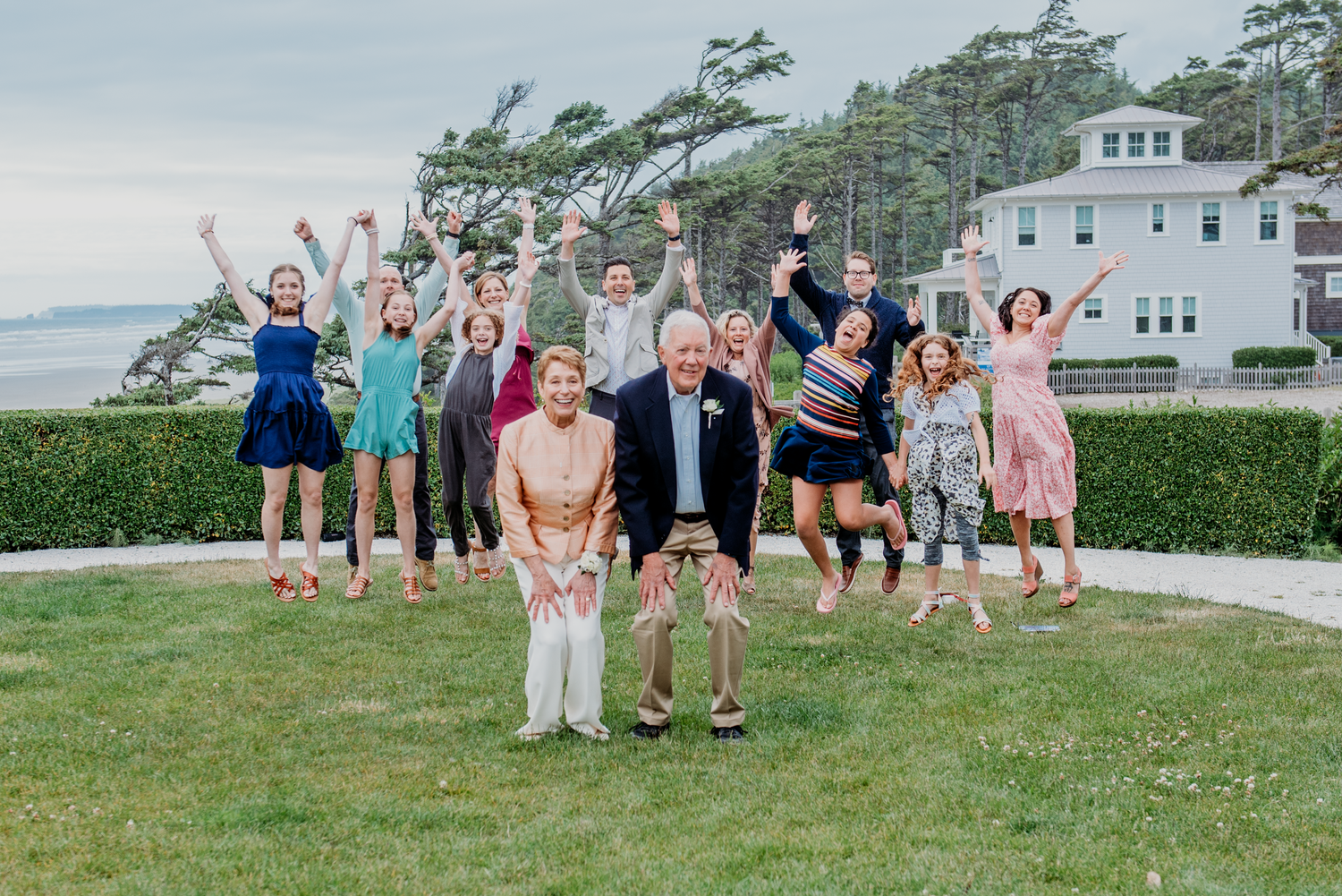 Large family group celebrates outdoors on green lawn, jumping joyfully with ocean view and house in background.
