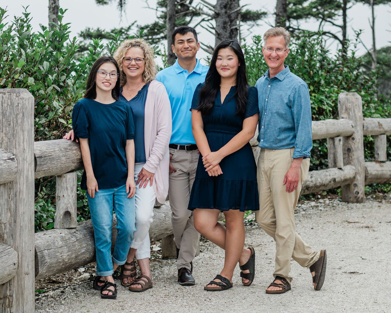 A smiling family of five poses outdoors near a wooden fence surrounded by lush greenery.