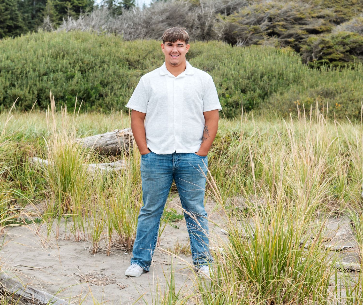 Person in casual clothing standing on sandy beach with grass and dunes in background.