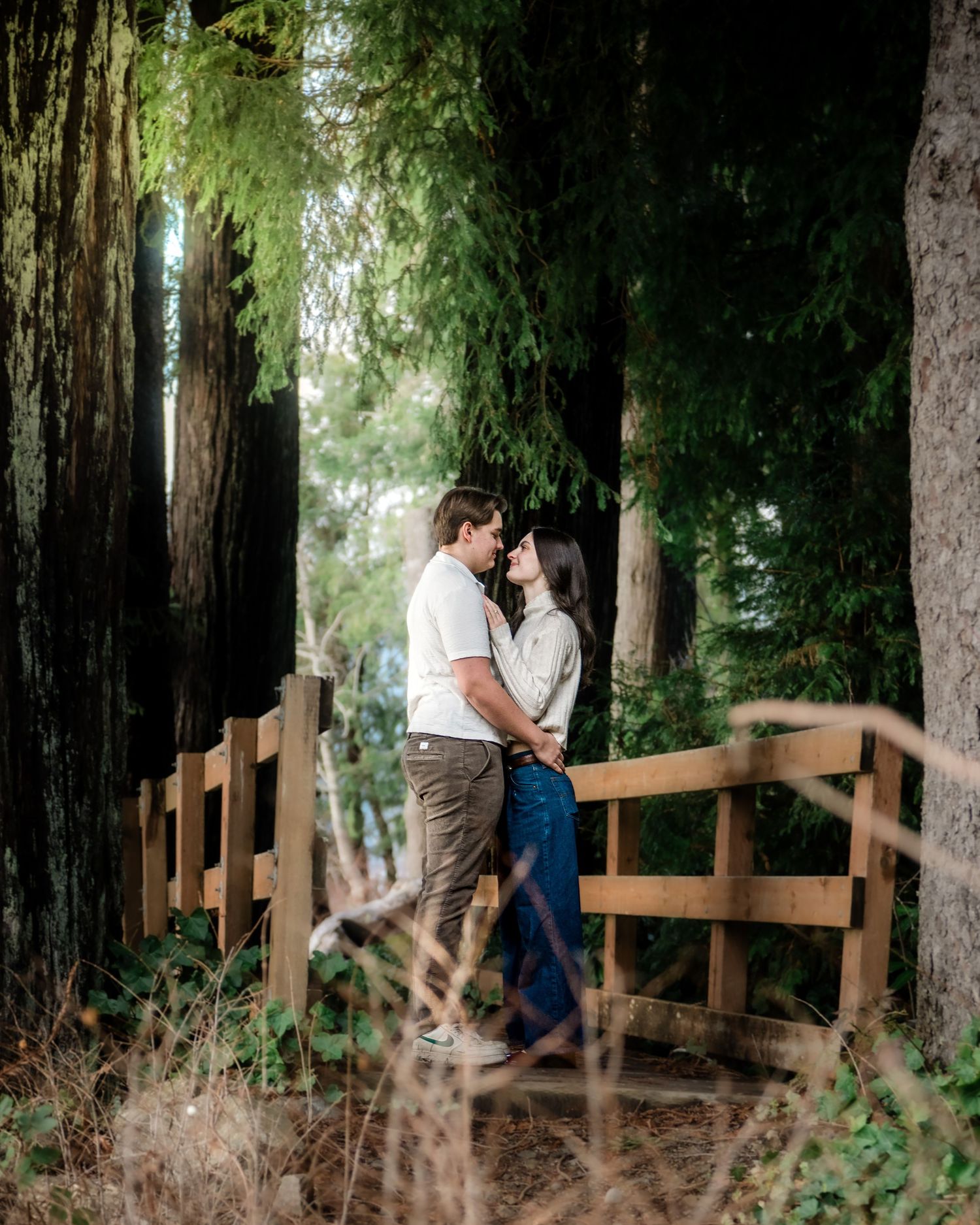 A couple shares an intimate moment on a rustic wooden bridge surrounded by towering redwood trees in a forest setting.