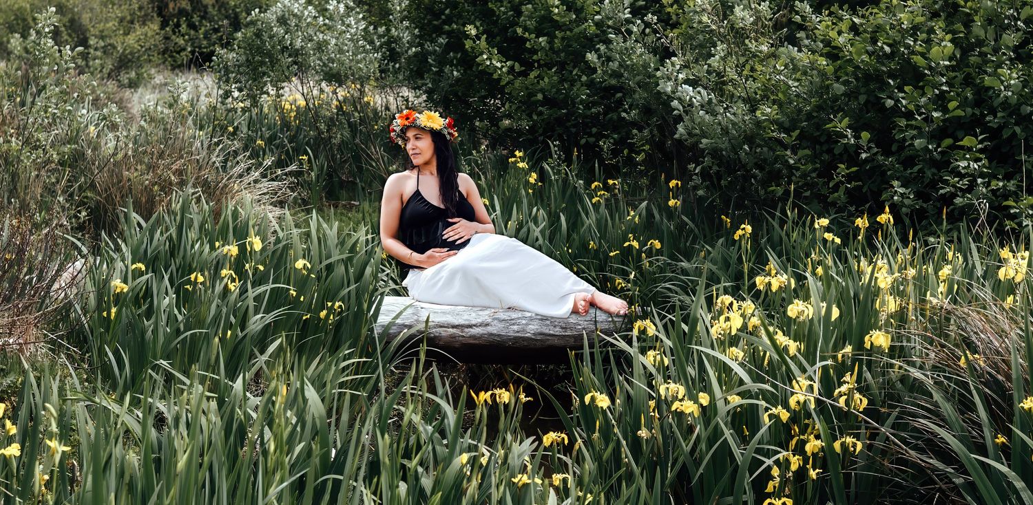 Woman in white dress and floral crown sits among yellow iris flowers in a lush green meadow.