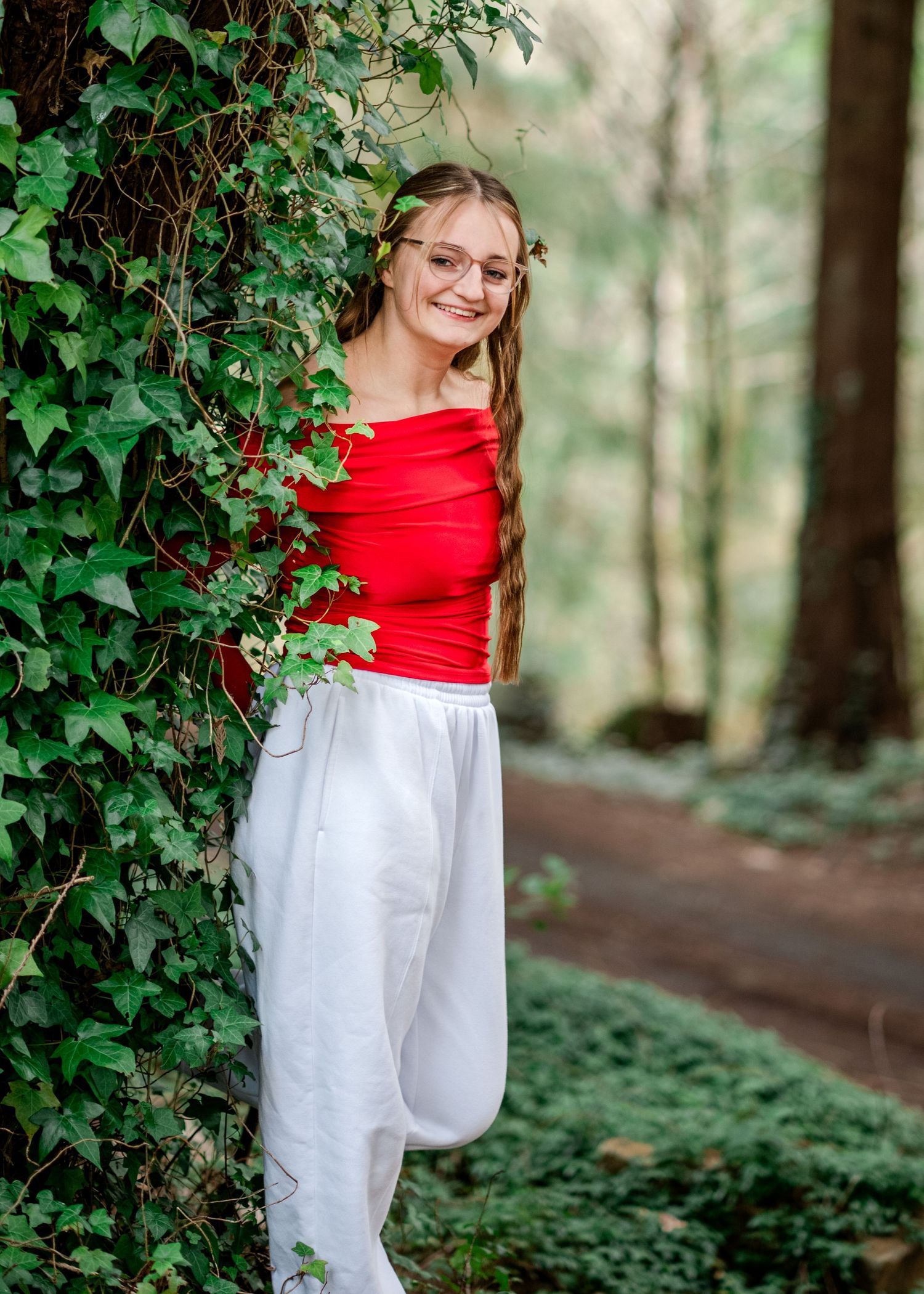 A person wearing a red sleeveless top and white flowy pants poses in a lush forest setting with green foliage.