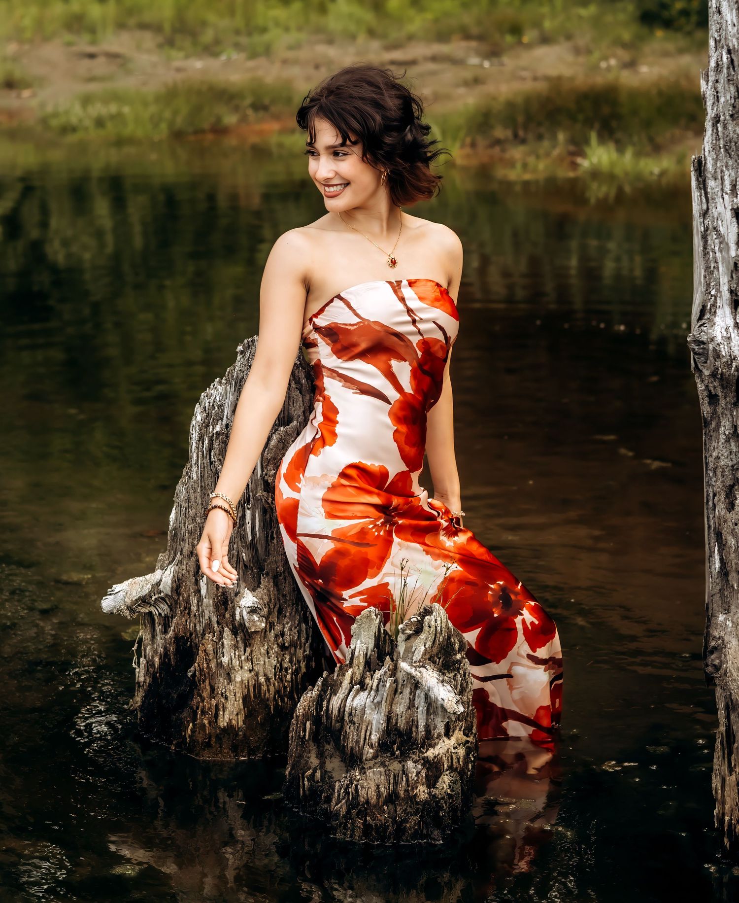 Woman in floral strapless dress posing on mossy rocks in a serene forest pond setting.